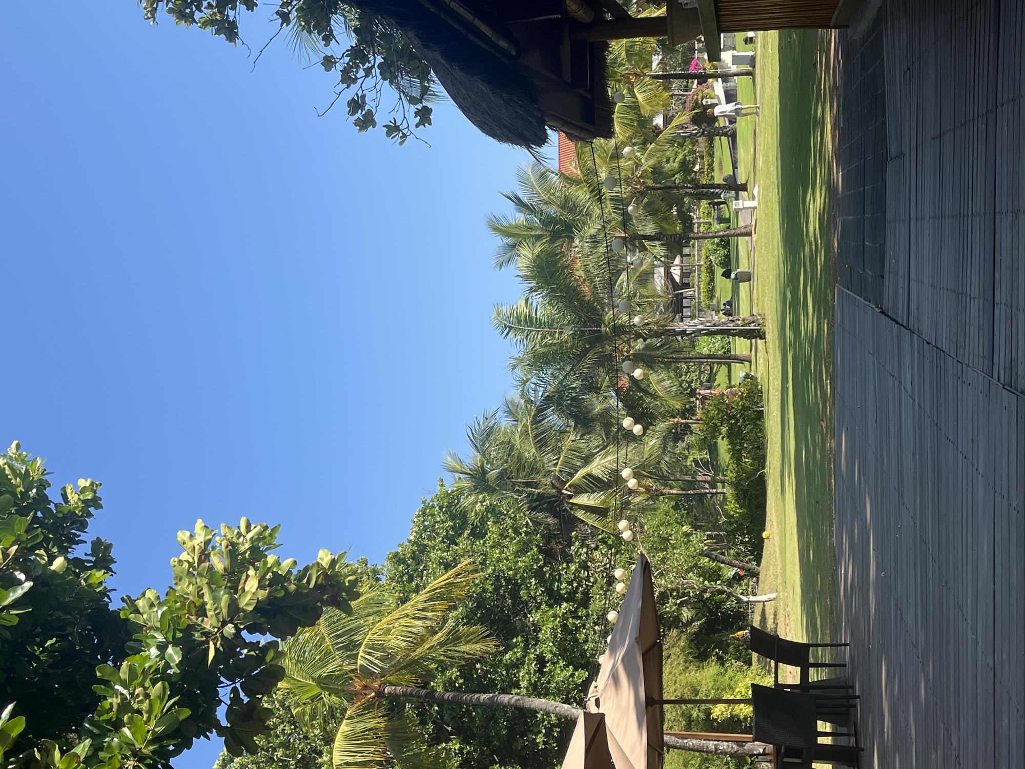 The image depicts an outdoor seating area at a resort in Kuta Selatan, Indonesia, on a clear, sunny day. In the foreground, a wooden deck is visible, with a table and chairs set up under a beige umbrella. Beyond the deck, a lush green lawn stretches out, dotted with numerous palm trees and other tropical foliage. In the background, several resort buildings with terracotta roofs can be seen, along with some distant figures enjoying the grounds. A thatched-roof structure, possibly a bar or open-air pavilion, is partially visible on the right side of the image. String lights are hung between some of the palm trees.