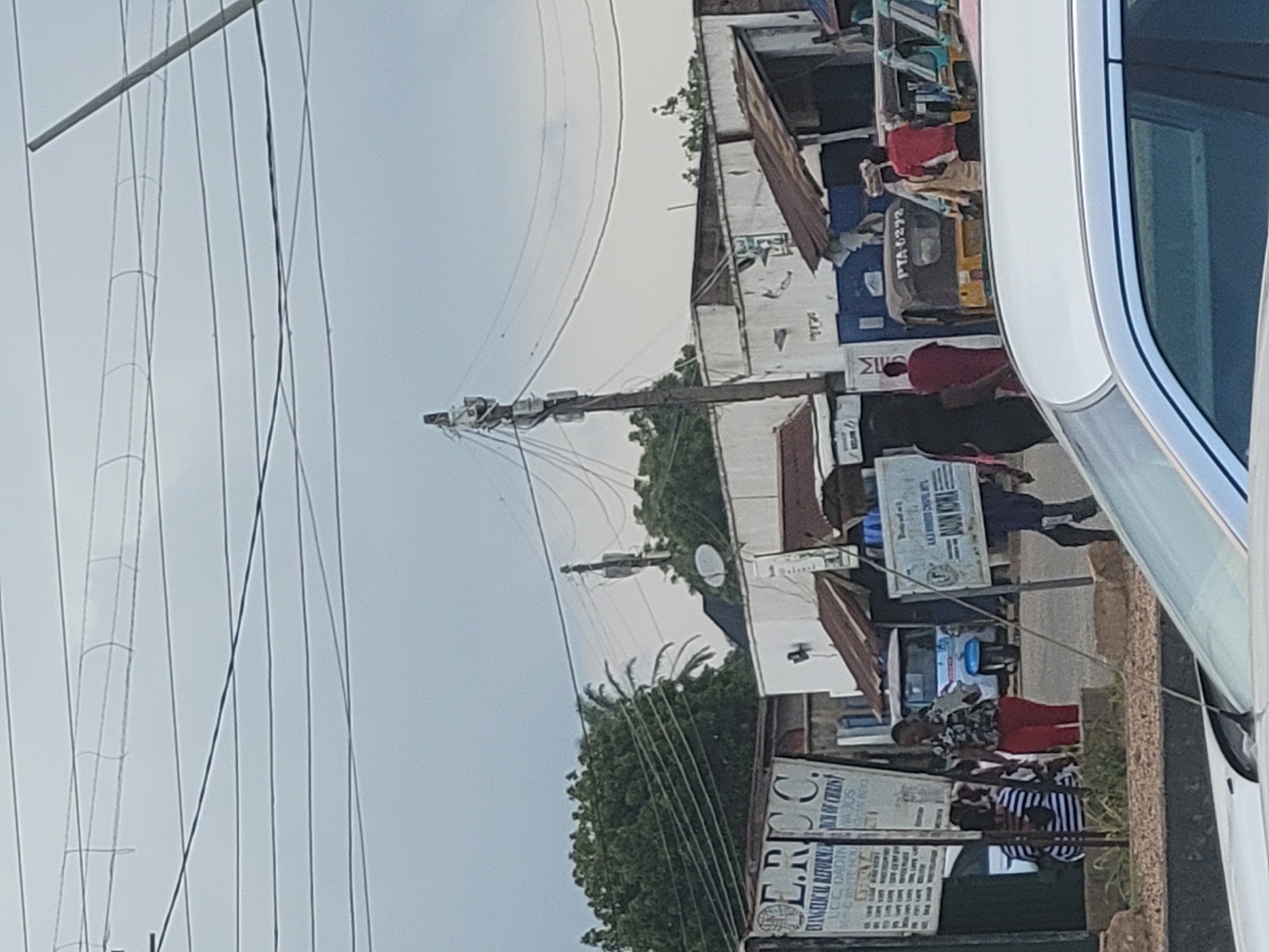 The image depicts a street scene, possibly in Africa, characterized by overhead power lines, a building with a tin roof, and several people. The scene appears to be during the day, with overcast skies. In the foreground, a white vehicle is partially visible on the right. A utility pole with electrical equipment is prominent in the middle ground. A yellow auto-rickshaw, identified by the license plate "PTA 6292", is parked behind the pole.
Several individuals are visible in the scene. One person in a red shirt is near the auto-rickshaw, while others are seen walking or standing near the buildings. A sign with text is partially visible, including "E.RIC C. EVANGELICAL REFORMERCH OF CHRIST" and "DADIN KOWA". The overall impression is of a bustling, everyday street in a developing area. The low-angle perspective and the presence of power lines suggest an urban or semi-urban environment. The weather appears to be cloudy, suggesting it might not be a bright, sunny day. The people's attire is casual, and there are no obvious signs of any specific event or emotional distress. The scene seems to capture a moment in daily life on this street.