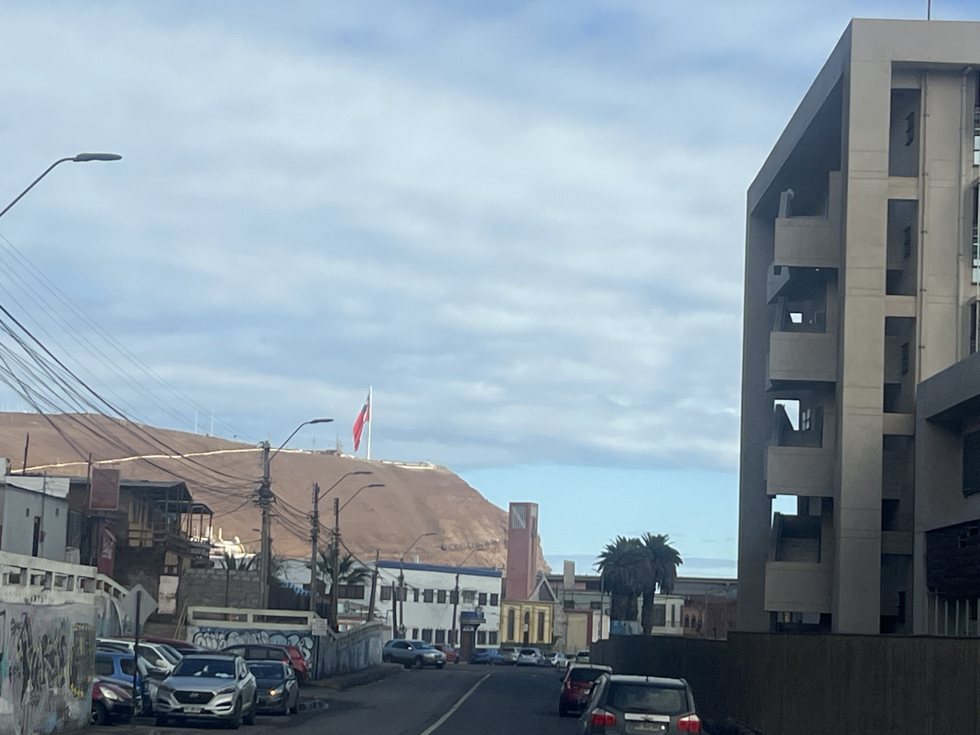 The image captures a street scene in Arica, Chile, dominated by a large, modern, multi-story building on the right side of the frame. This building appears to be made of concrete and features numerous balconies and open window spaces, suggesting a potentially unfinished or architecturally distinct structure.
The street in the foreground is lined with parked cars and shows signs of urban wear, including graffiti on a low wall to the left. Several streetlights are visible along the road.
In the background, a prominent red-brown hill rises, atop which a Chilean flag is flying from a tall flagpole. Below the hill, a cluster of buildings forms a townscape, including a distinctive red-and-yellow tower that resembles a clock tower or lighthouse. Palm trees are also present in the background, typical of coastal regions. The sky is overcast with scattered clouds. The overall impression is of a coastal city with a mix of modern architecture, older structures, and natural topography.