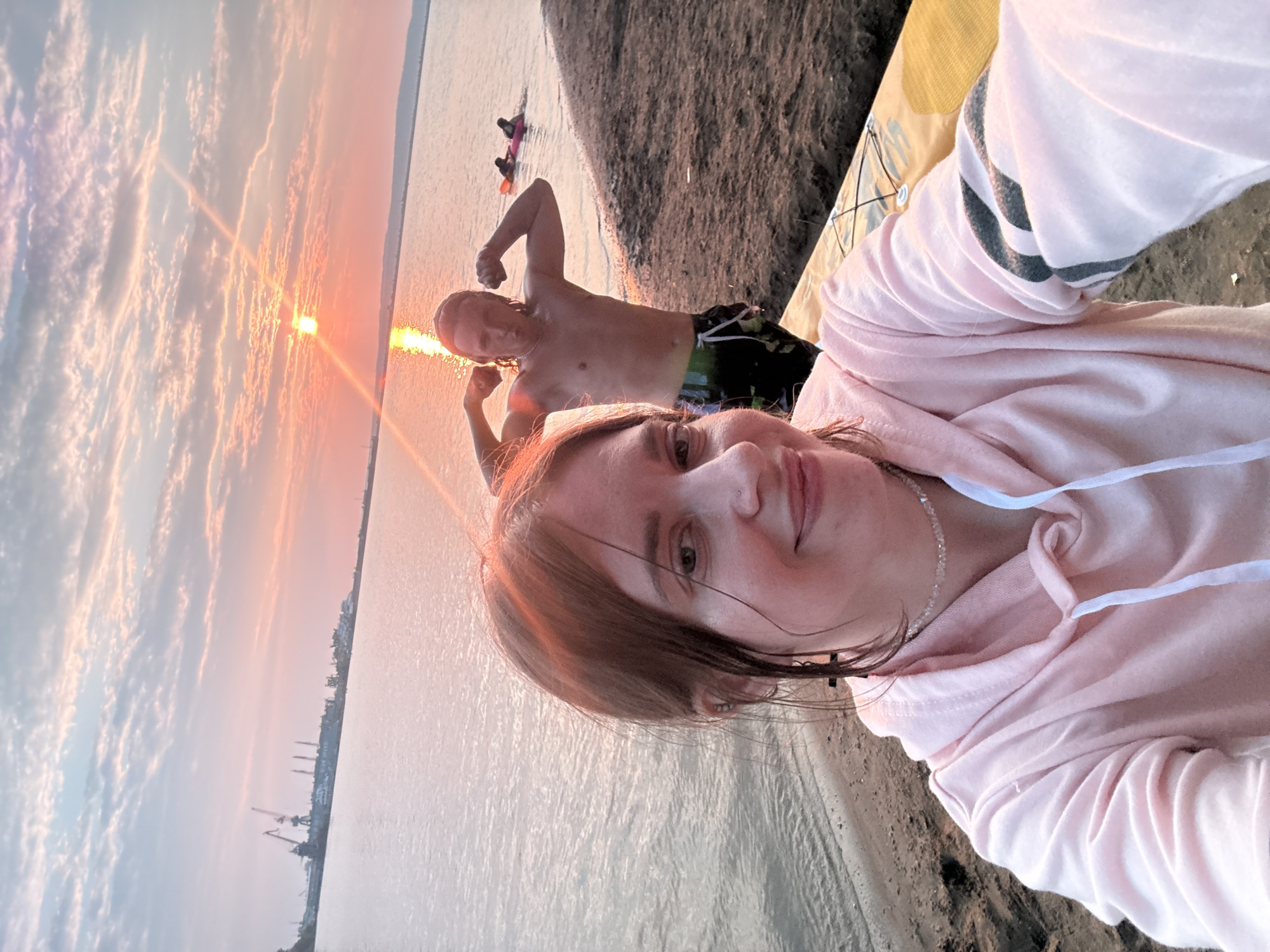 In an outdoor selfie, a woman with brown hair, wearing a pink hoodie and a clear necklace, smiles at the camera. Behind her, a man with light brown hair, shirtless and wearing patterned swim trunks, flexes his biceps. The scene is set at a beach with water in the background, where two people are floating on inflatable toys. Cranes are visible in the distance on the horizon, suggesting an industrial area. The sky is illuminated by a vibrant sunset.