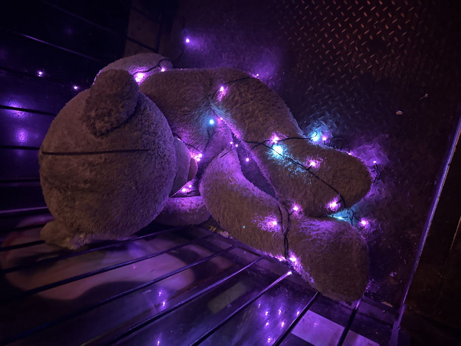 A large, greyish-brown plush teddy bear is situated in a dimly lit environment. The bear is seated, slumped forward with its head down, on a dark, textured metal diamond plate floor. A string of small LED lights, predominantly purple with interspersed blue lights, is wrapped around the bear's body, providing the primary illumination for the scene. To the left of the bear, dark, vertical metal bars or slats are visible, behind which a faint, lighter-colored surface can be discerned. The scene is static, devoid of human presence or active interactions. This location is within the city of Tel-Aviv, Israel.