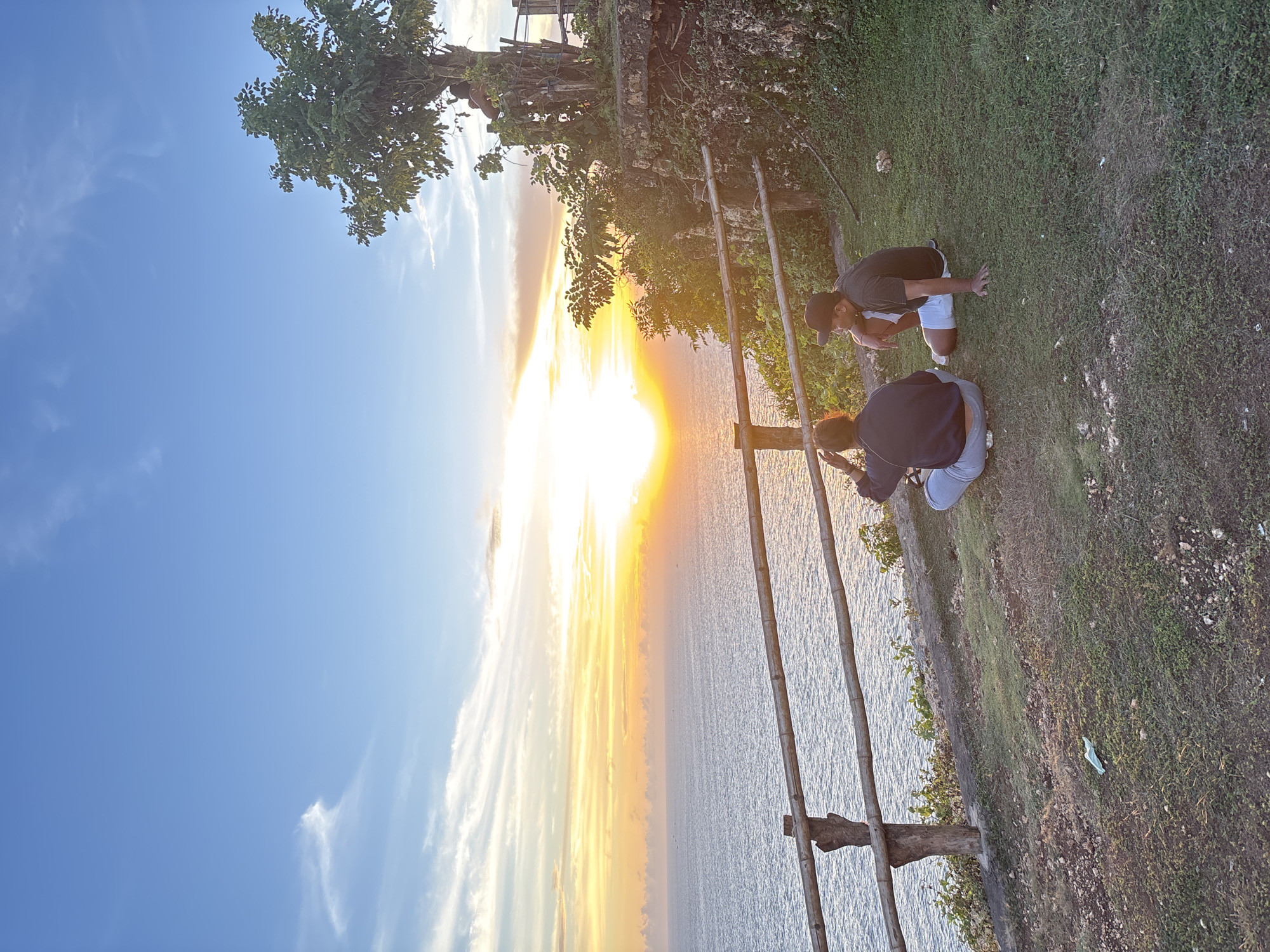 The photograph captures a scenic view at a location in Kuta Selatan, Indonesia. The setting appears to be on a cliff overlooking the ocean during sunset. The sky transitions from blue to shades of orange and yellow as the sun sets over the water, creating a vibrant display.
Two individuals are present on the grassy clifftop. One person is wearing a dark shirt, light shorts, and a baseball cap. The other individual is dressed in a dark jacket and gray pants. Both figures are kneeling and seem to be interacting near a simple bamboo structure.
A makeshift bamboo ladder or railing is constructed near the edge of the cliff. The vegetation on the cliff includes trees and grass.
