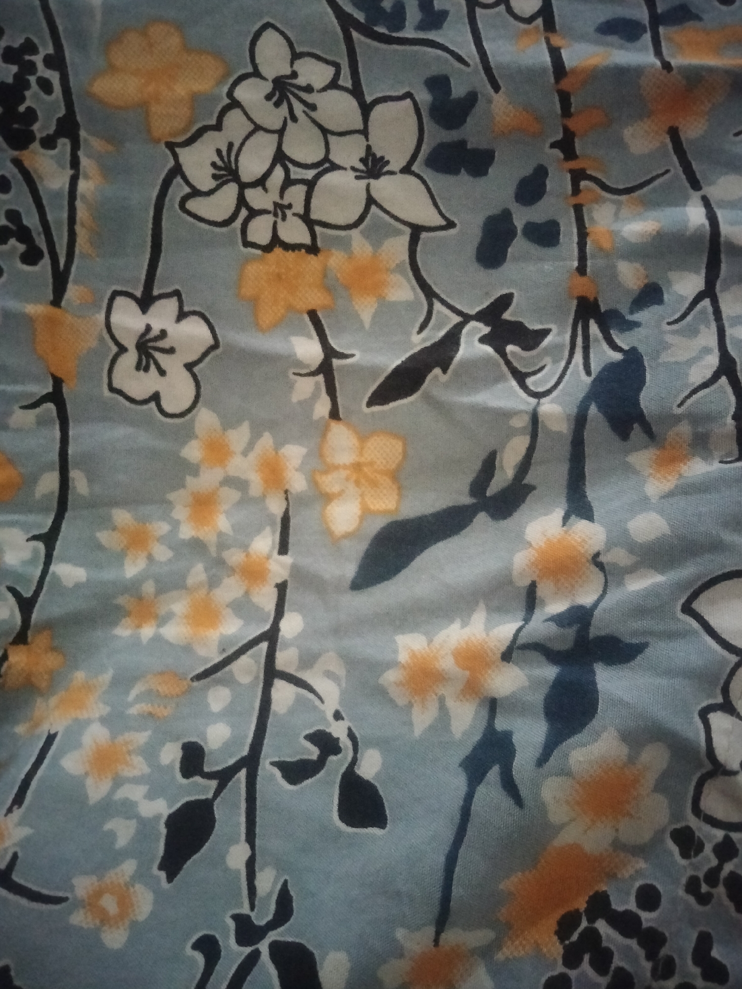 The image is a close-up, top-down view of a floral patterned fabric with a light blue background. The pattern consists of stylized flowers and leaves. The flowers are depicted in white and orange, with black outlines for some. There are also dark blue leaf shapes. The fabric appears to be slightly wrinkled, suggesting it might be draped or folded. The image is a detail shot and does not contain any elements that would suggest its location within the city of Suko, Indonesia.
