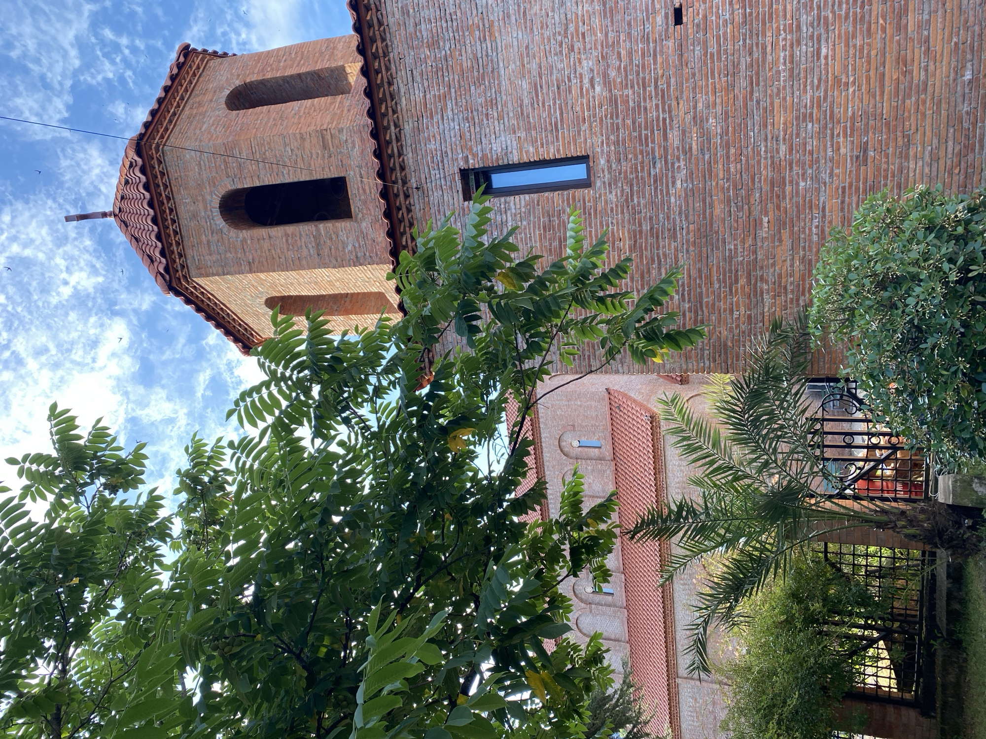 A red brick church with a tiled roof stands under a blue sky with scattered clouds. A tall, square bell tower with arched openings and a cross on top is visible on the right side of the image. A section of the main church building with arched windows and a red tiled roof is visible on the left. Lush green trees and a palm tree partially obscure the view of the church. The scene is likely in Batumi, Georgia, given the architectural style and the presence of sub-tropical vegetation.
