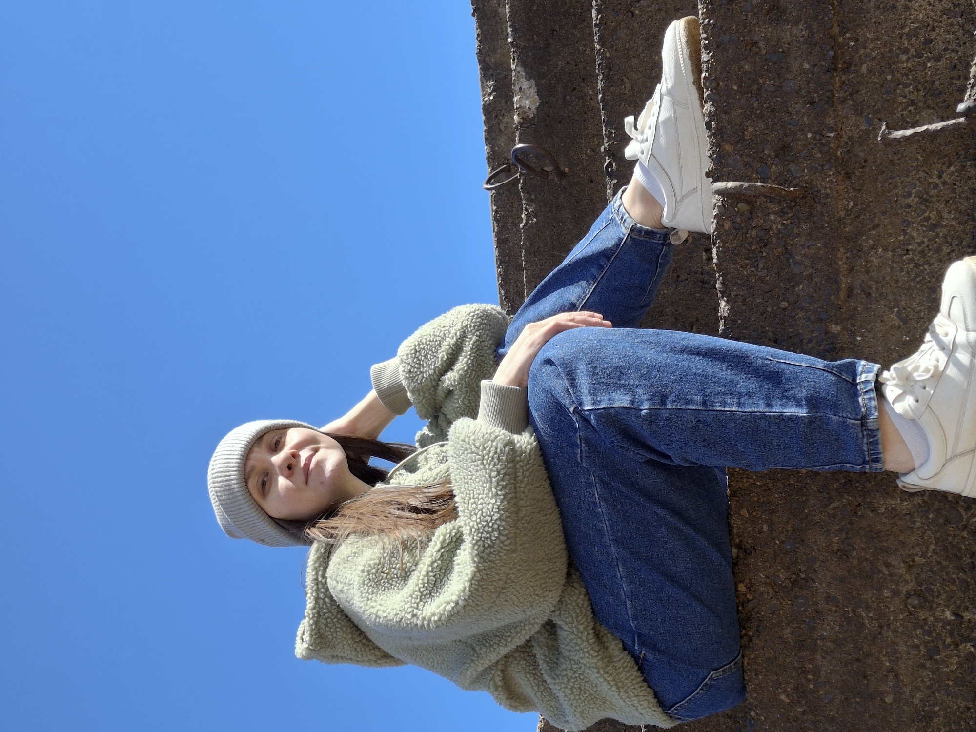 This vertical image, taken from a low angle, features a young woman posed against a clear blue sky and a textured concrete surface. The woman is positioned along the right side of the frame, appearing to lie or lean on the rough structure.
The subject is a young woman with light brown hair, wearing a light grey ribbed beanie, a light green fuzzy hooded jacket, blue denim jeans, and white sneakers with white socks. She looks towards the camera with a calm, slightly smiling expression. Her right hand is near her head, while her left hand rests casually on her left thigh.
The background on the left is a vibrant, clear blue sky, indicating bright, sunny weather and strong daylight, likely midday. The right and bottom portions of the image reveal a dark, coarse concrete or stone surface with visible aggregate and some embedded metal hooks or rebar. This surface suggests an architectural element like a wall, pillar, or large block.
The activity is a casual portrait or lifestyle photo, with the woman striking a relaxed pose. There are no explicit location cues within the image itself beyond the general urban/architectural features, but the provided context states the location is Chaikovsky, Russia. No visible text is present in the image.