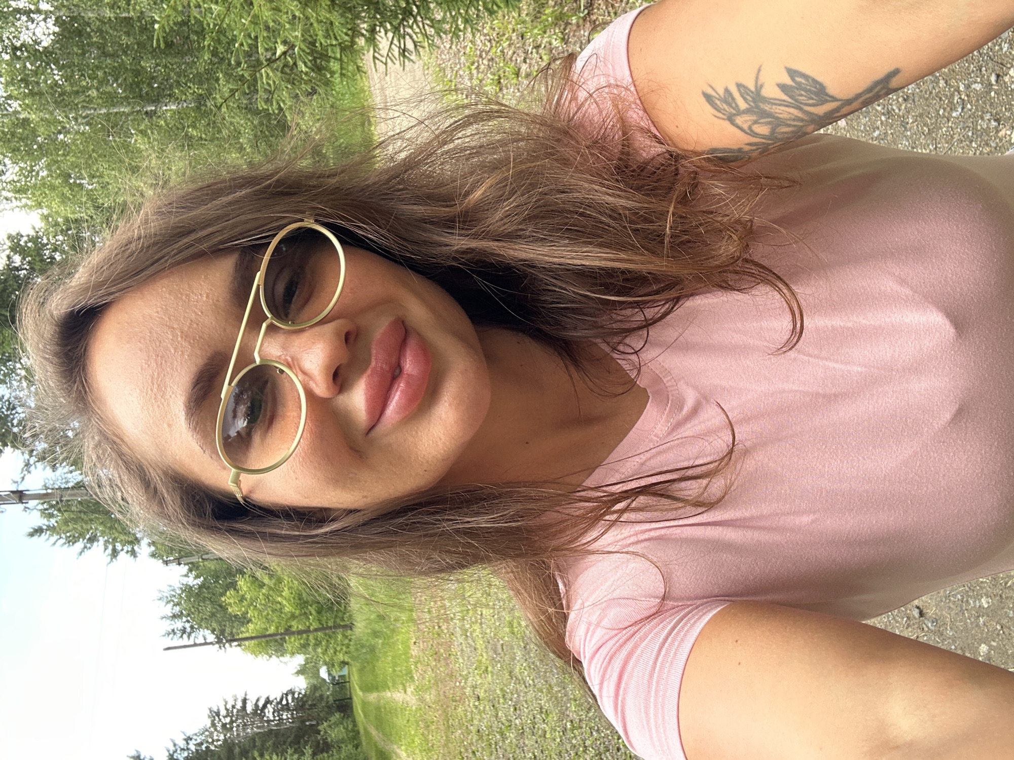 A woman is taking a selfie outdoors, with a dirt road and trees in the background. She is wearing a pink t-shirt and gold-rimmed glasses. Her hair is long and brown, and she has a tattoo on her left arm. The location is identified as Duvankul', Russia. The weather appears to be sunny and warm, with a clear sky.