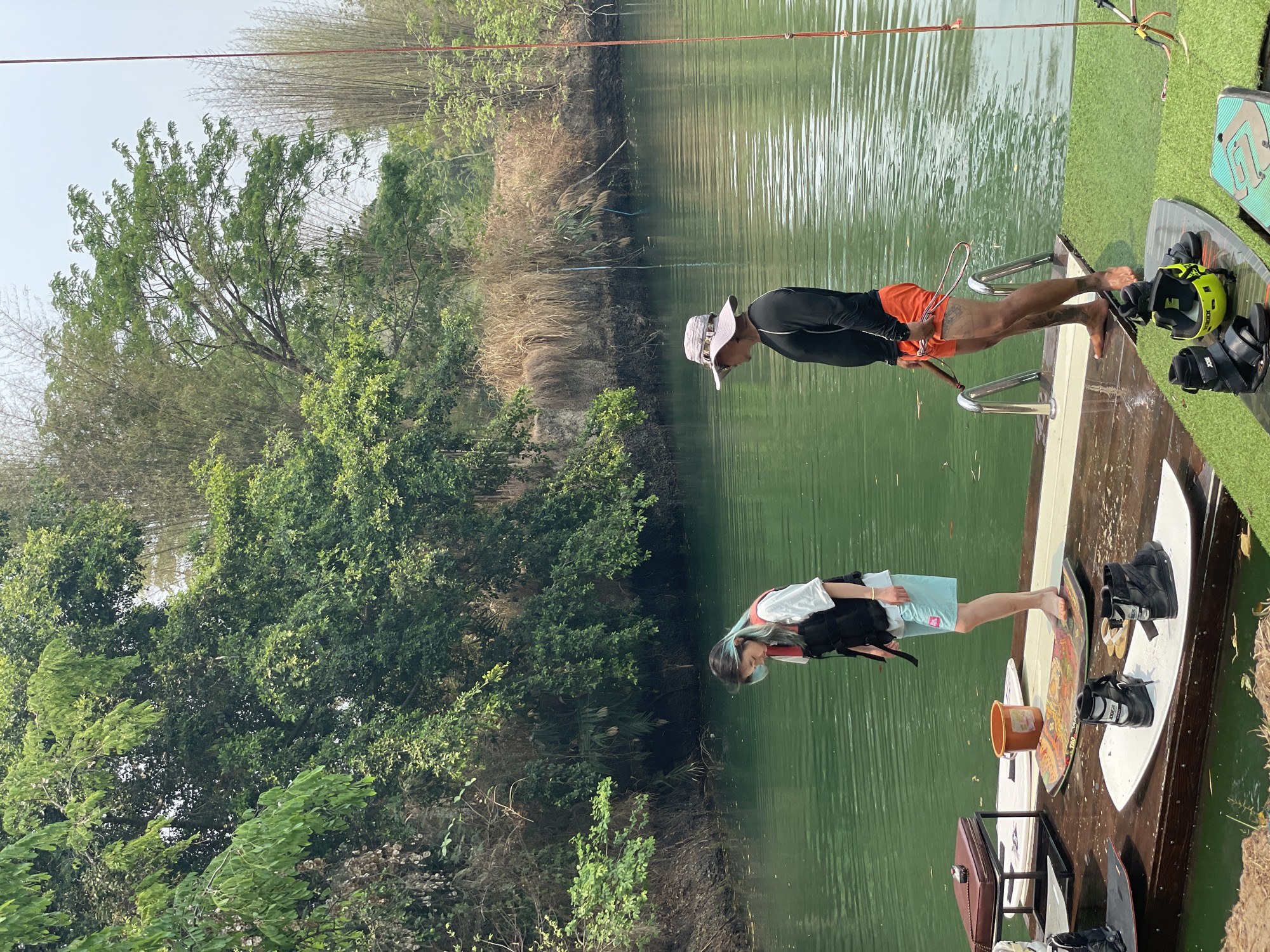 Two individuals are on a wooden dock at a lake, likely preparing for water sports. A man in a black long-sleeved shirt, orange shorts, and a sun hat stands near the water's edge, with a metal ladder extending into the water. Next to him is a wakeboard and bindings. A younger person with dyed blue hair, wearing a white t-shirt and blue shorts, stands further back on the dock. A life vest lies next to the individual. Various wakeboards and accessories, including bindings and an orange bucket, are also on the dock. The background consists of lush green trees and reeds growing along the edge of the lake. The location is Tambon Nam Phrae, Thailand.