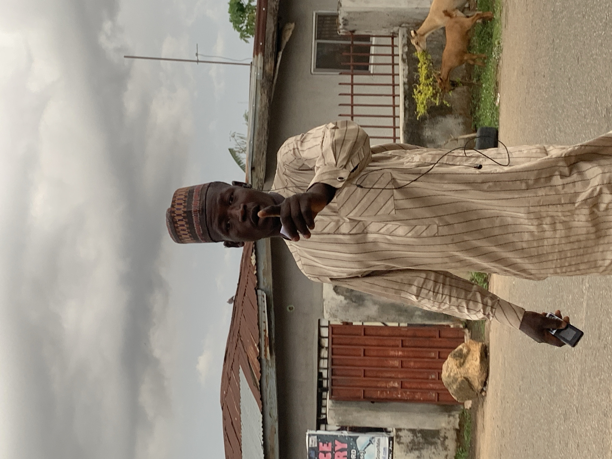 A middle-aged man wearing a traditional embroidered cap and a striped, long-sleeved garment stands on a paved road. He holds a dark object to his mouth with his right hand and a cell phone in his left. A small microphone is clipped to his shirt, with a wire extending to his ear.
Behind him, a light-colored building with a partially rusted metal roof and a window with iron bars is visible. A reddish-brown gate is beside the building. Further back, two goats are grazing on a patch of greenery.
A poster or sign is partially visible at the bottom left of the frame. The sky is overcast with grey clouds. The scene is characteristic of Jos, Nigeria, with its combination of modern technology and traditional attire within an urban environment.