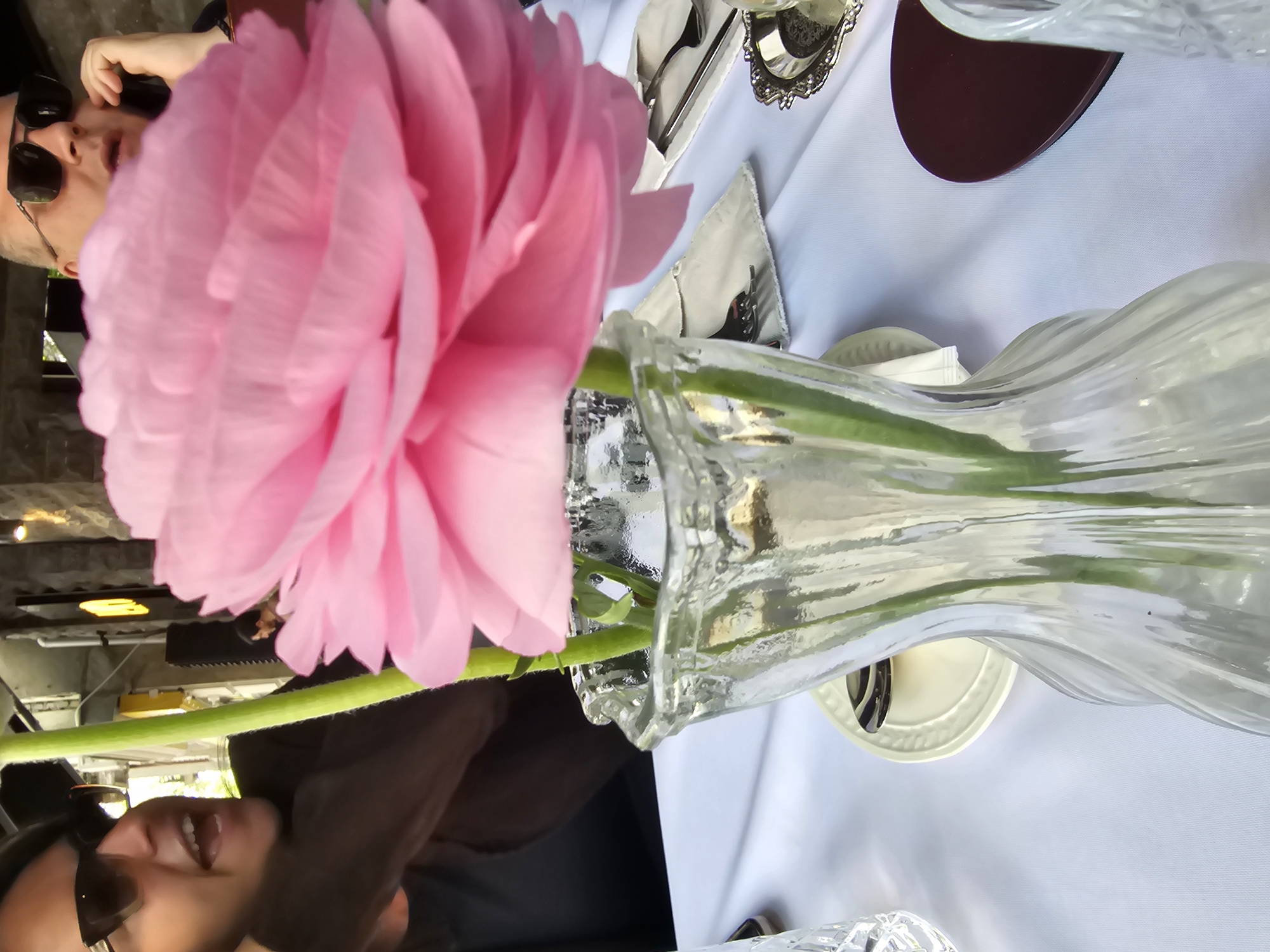 A close-up shot shows a pink ranunculus flower in a clear glass vase on a table with a white tablecloth. Silverware and a napkin are visible nearby, suggesting a table setting. The shot is taken outside, possibly in a restaurant or cafe, given the blurred background that indicates a public space. Parts of two people are visible: one in the background wearing sunglasses and one in the foreground also wearing sunglasses. Given the style of the building in the background, the location may be in Tbilisi, Georgia, which is a city known for its open-air restaurants and distinctive architecture.