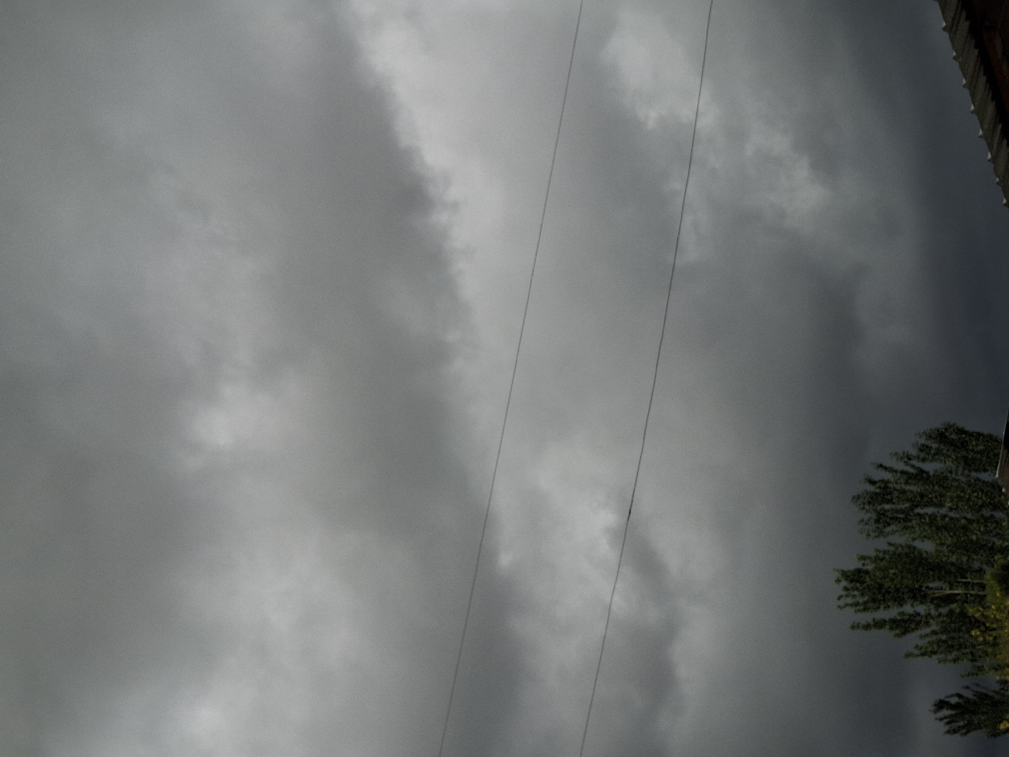 A sky filled with dark, brooding clouds hangs over a scene in Jeti-Ögüz, Kyrgyzstan. Two thin, dark wires stretch horizontally across the middle ground, suggesting the presence of utility infrastructure. Below these wires, the tops of lush green trees are visible, their foliage dense and vibrant against the somber sky. The uppermost portion of a dark, corrugated roof juts into the frame from the bottom right corner, hinting at a residential or utility structure. The overall impression is one of impending weather, with the heavy clouds dominating the composition.