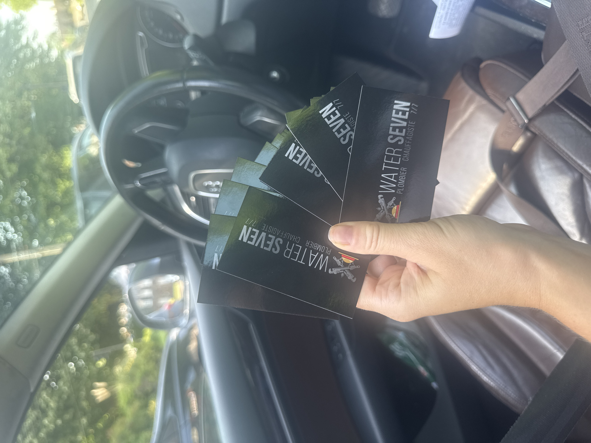 The scene is an interior shot of a car, presumably in Marcq-en-Barœul, France, based on the business cards held by a hand in the foreground. The hand holds several business cards for "WATER SEVEN," a plumbing and heating company ("PLOMBIER CHAUFFAGISTE 7/7"). The car's steering wheel and dashboard are visible in the background, suggesting the photo was taken from the driver's seat. The car seat and seatbelt are also visible. Outdoor bokeh light spots suggests the presence of greenery.