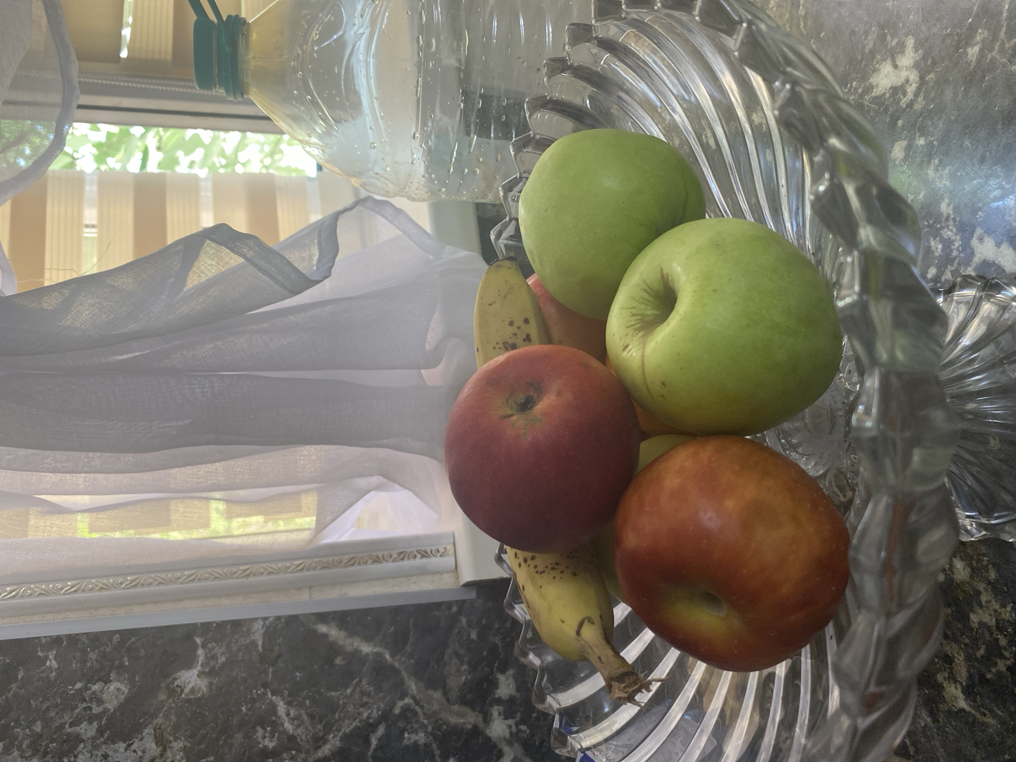 In Alkhan-Kala, Russia, a crystal fruit bowl sits on a dark countertop near a window. The bowl contains two red apples, two green apples, and one partially visible banana. Behind the bowl, a large clear plastic bottle filled with water stands to the right, and sheer white curtains hang in front of the window. The window blinds are partially open, allowing light to filter through, and a glimpse of green foliage is visible outside. The countertop has a marbled pattern.