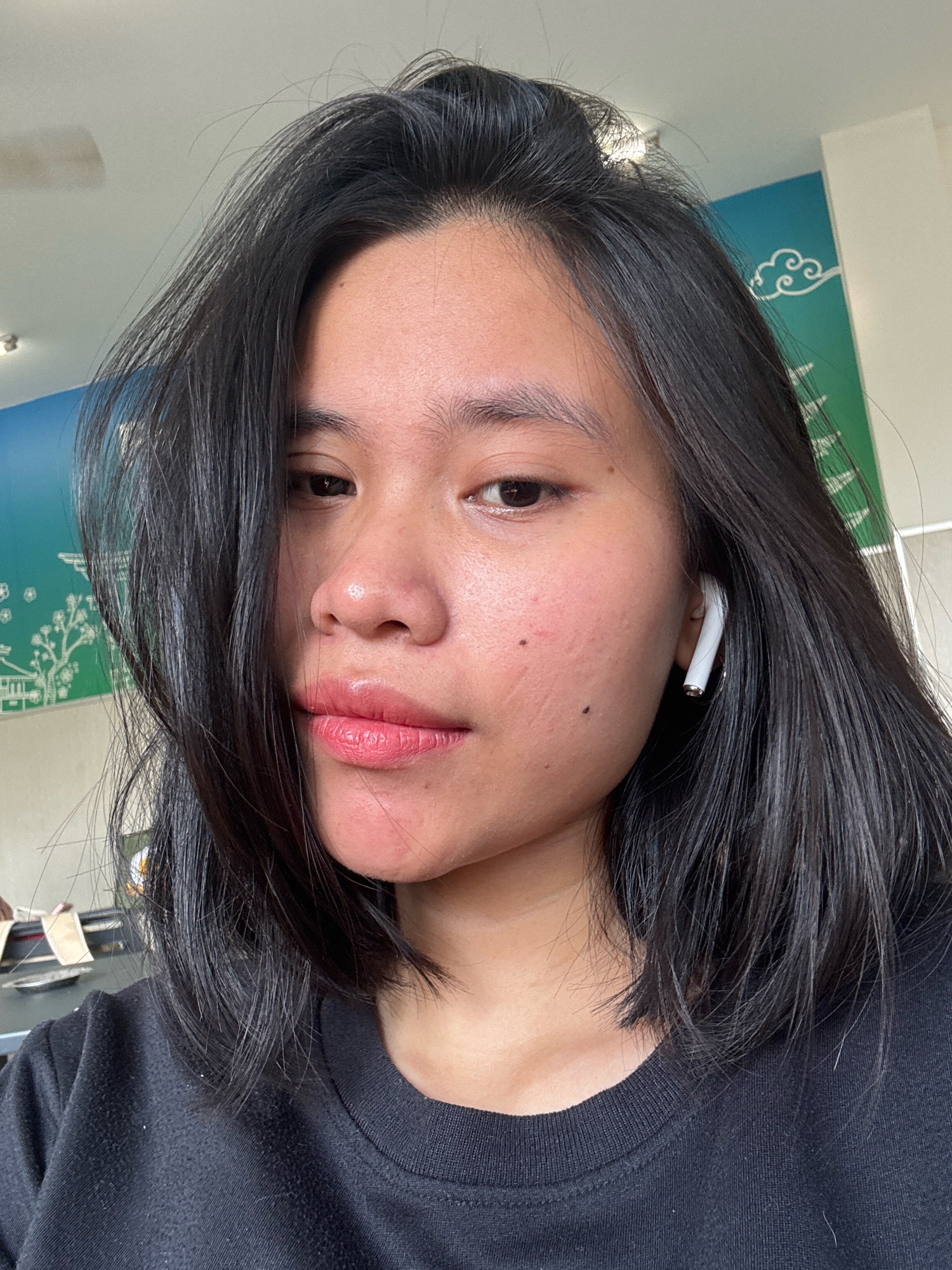 A close-up selfie shows a young woman from the chest up. She has shoulder-length dark hair, fair skin with some redness and blemishes on her cheeks, and is wearing a black t-shirt. She has on a light pink lipstick. A white wireless earbud is visible in her right ear. The background appears to be indoors with a wall featuring a green and blue mural with white cloud and tree designs. The image is likely taken in East Jakarta, Indonesia.