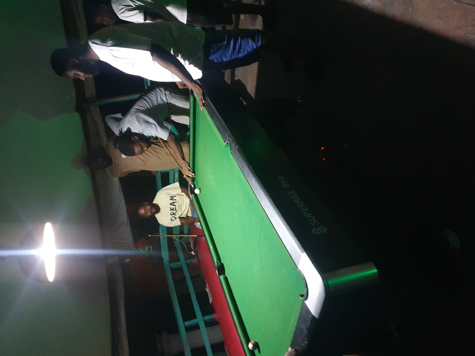 An outdoor setting at night, possibly a bar or lounge, in Lokoja, Nigeria, features a group of men playing pool. A green pool table with white and red balls is the central focus. The lighting is dim, originating from an overhead lamp. One man in a white t-shirt and blue shorts is actively playing, aiming a cue stick at a ball. Another man in a striped shirt is also positioned near the table, appearing to be involved in the game. A third man in a tan shirt stands behind the player, observing. A fourth man in a yellow t-shirt with "DREAM" printed on it sits on a bench or railing to the left of the table. The background consists of a railing, a dark wall, and some foliage. The overall atmosphere is casual and social.