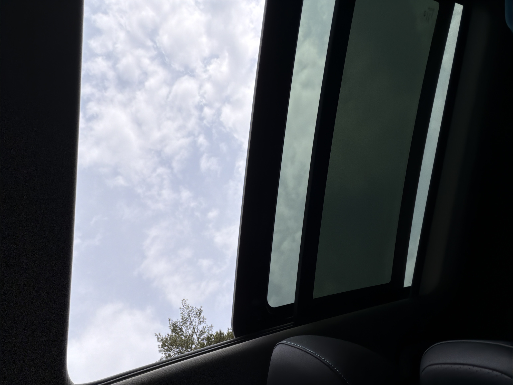 The image shows a view through an open sunroof in a car, looking upwards at a cloudy sky. Part of a tree is visible through the opening. The interior of the car is dark, showing part of a seat in the lower right corner. The scene appears to be an ordinary moment captured from inside a vehicle. There is no indication of a specific location within Chaikovsky, Russia, or any other identifying details in the image.