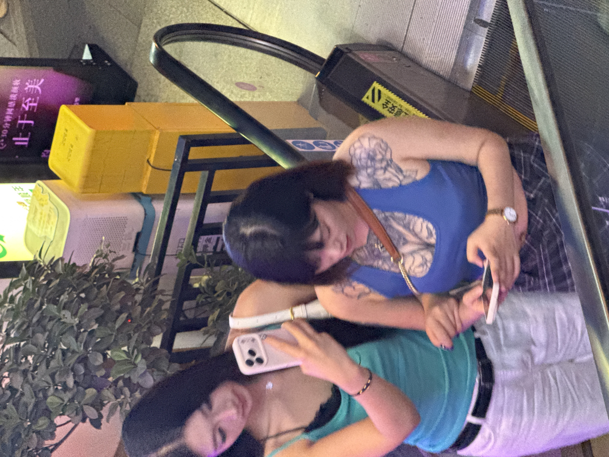 In a commercial setting in Liangjiang, China, two young women are on an escalator. The woman in the foreground, on the right, has dark hair and is wearing a blue tank top with a large tattoo on her upper chest and shoulder. She is looking down at her phone, which she is holding in her right hand. She has a watch on her left wrist. The woman on the left has long dark hair and is wearing a turquoise tank top and white pants. She is holding a smartphone in her right hand, seemingly taking a photo or video. She has a thin bracelet on her left wrist. The escalator has yellow and black safety strips. In the background, there is a plant, yellow boxes, and a poster with Chinese text.