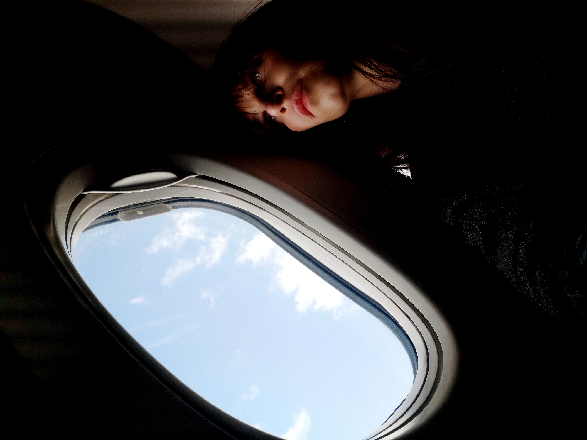 A low-angle, indoor shot captures a woman looking down toward an airplane window. The window is open, revealing a light blue sky with a few white clouds. The woman has dark hair and fair skin, and is partially obscured by shadow. The scene is characterized by strong contrast and is mostly dark, except for the face of the woman and the sky visible through the window. The location is not evident based on the image.