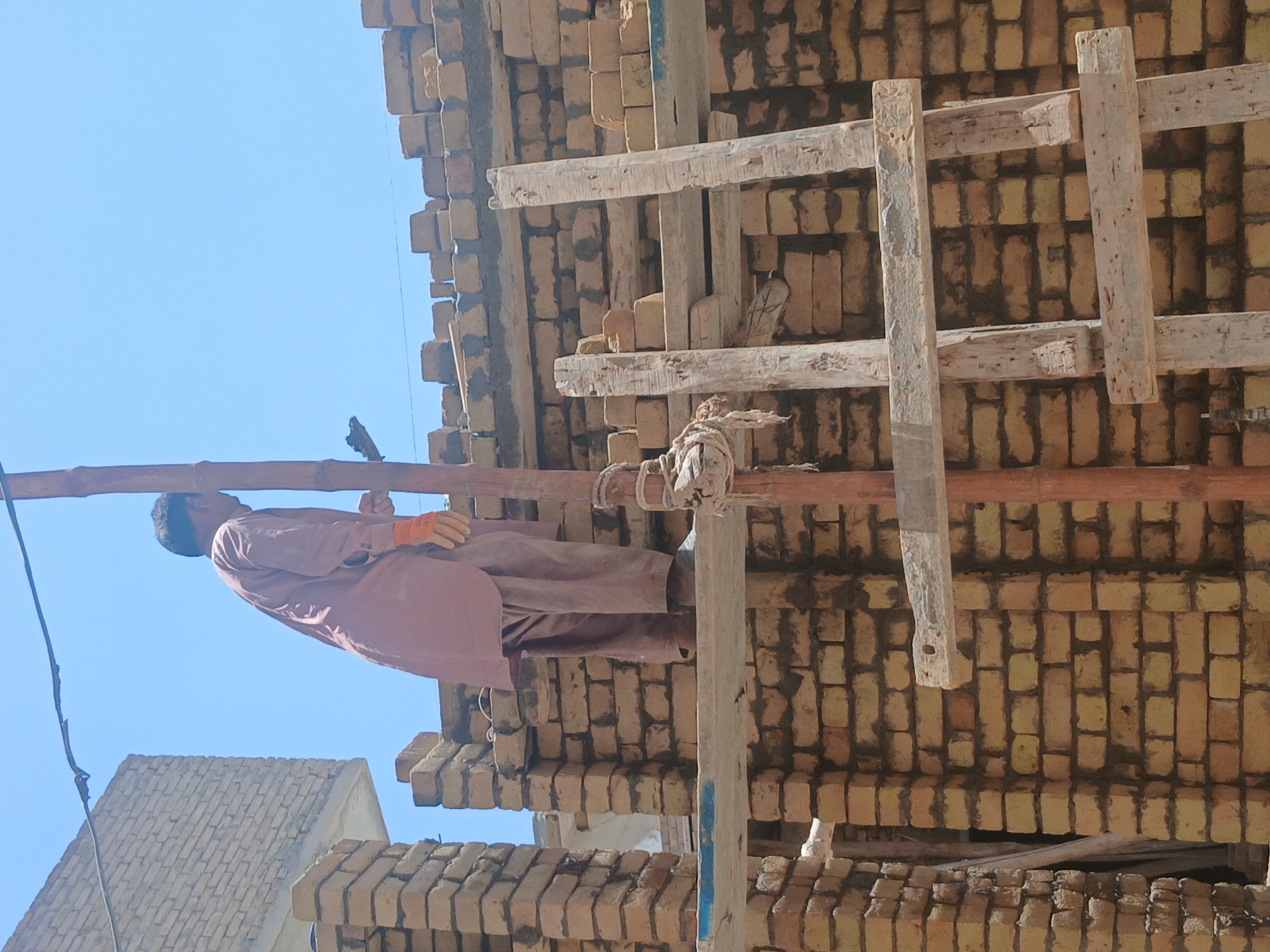 A man in a light brown tunic and pants, wearing orange gloves, is working on a brick structure under construction. He is standing on a makeshift bamboo scaffolding. The structure is made of yellow-brown bricks. The scene is set against a clear blue sky and other buildings are visible in the background. The location is in Quetta City Tehsil, Pakistan.