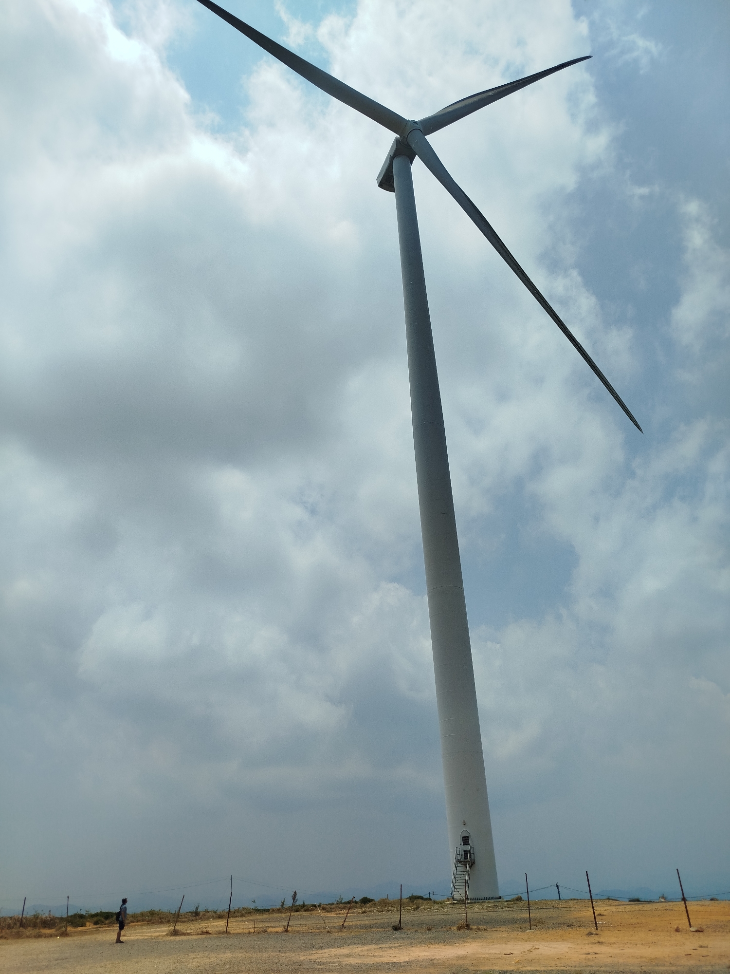 A ground-level, wide-angle shot shows a wind turbine against a cloudy sky, located in what is described as the city of Phát Chi, Vietnam. The turbine has three blades, two of which are prominently visible. The turbine's tower is white or light gray. At the base of the tower, there is a door or entrance with a ladder-like structure leading to it. A fence, constructed from vertical poles connected by wires, runs along the perimeter of the area. In the lower left corner of the image, a person stands facing the turbine. The ground is dry, covered in gravel and sparse vegetation. In the distance, a low mountain range can be discerned. The sky above is filled with a mix of light and dark clouds, suggesting an overcast day.