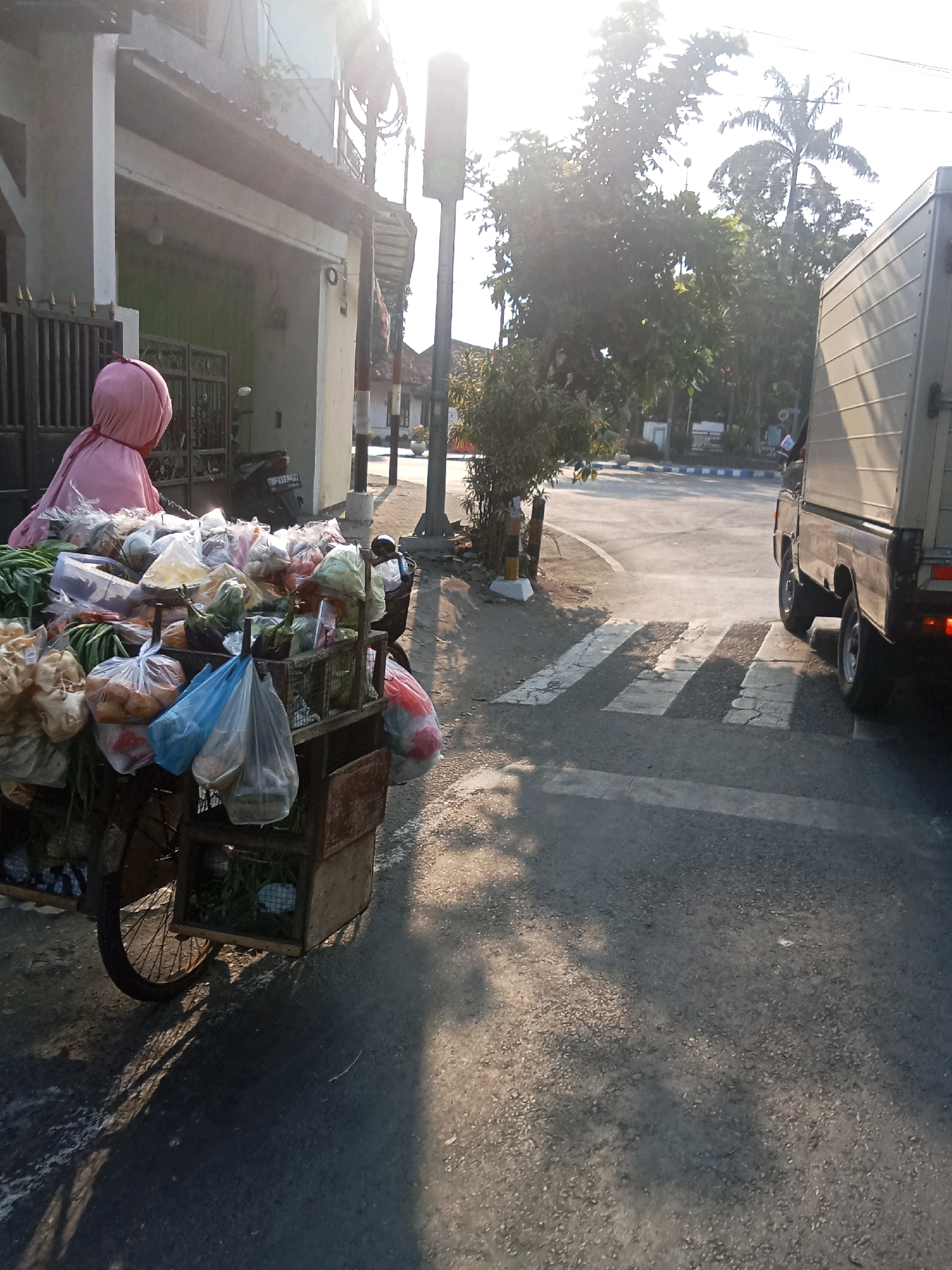 A vendor, wearing a pink hijab, pushes a loaded bicycle cart along a street in Jombang, Indonesia. The cart is filled with various fresh produce, including watermelons, vegetables, and bagged goods, suggesting the vendor is on their daily route. The scene captures a typical urban street with buildings lining one side and a road with a crosswalk on the other. A light-colored truck is visible on the road, stopped near the crosswalk. The image is taken during daylight hours, with the sun casting long shadows, indicating it's likely early morning or late afternoon. The presence of a traffic light and palm trees further contributes to the Indonesian urban setting. The overall atmosphere is that of daily commerce and movement within the city.