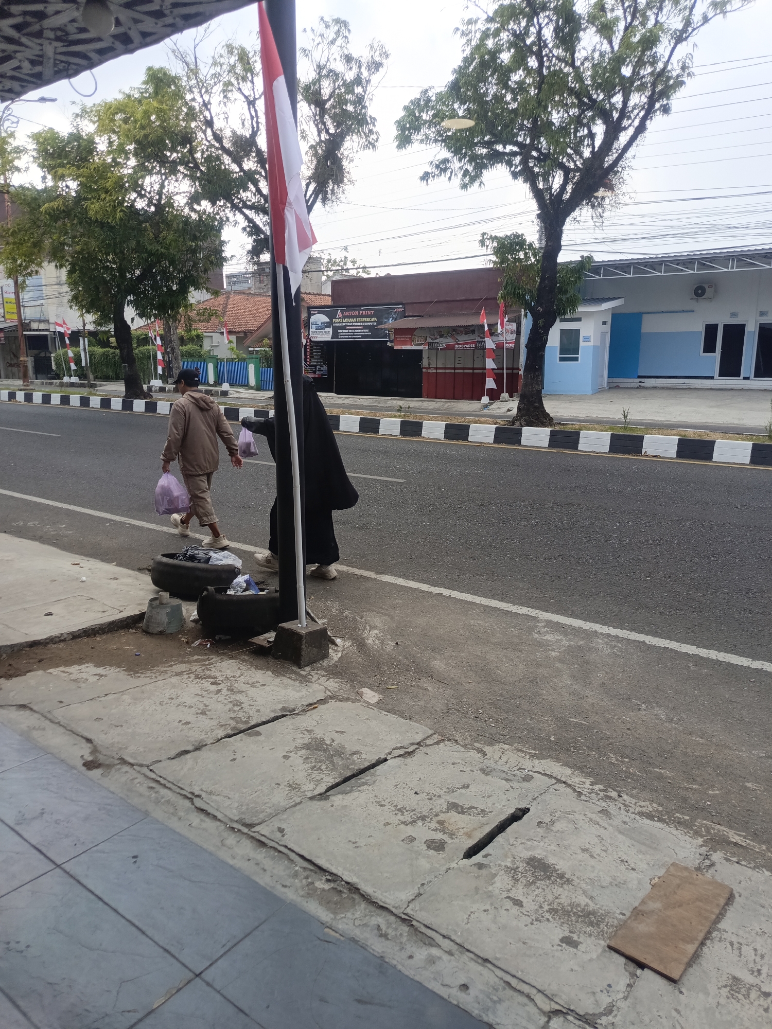 In Desa Gunungsimping, Indonesia, a man and a woman are walking on a sidewalk next to a road. The man, wearing a light brown hooded jacket and matching pants, carries two purple plastic bags. The woman, dressed in a black burqa, also carries two purple plastic bags. In front of them, two discarded tire rims are placed on the sidewalk, filled with debris. Behind the people, a red and white Indonesian flag stands tall on a pole. The road features a white and black striped border. On the right side of the road, a building with a blue and white facade is visible, with an air conditioning unit mounted on its exterior. The sky is overcast.
