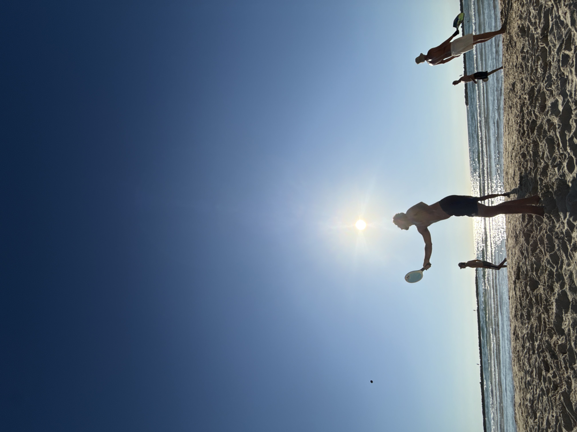 The image captures a group of people playing beach tennis on a sandy shore with the ocean in the background. The sun is bright and positioned in the sky, creating a slight glare. Several individuals are visible, silhouetted against the sky, some holding tennis rackets and appearing to be in the midst of a game. The scene is set on a beach, likely in Tel-Aviv, Israel, given the typical coastal environment and recreational activity. The sand appears dry and textured, with waves breaking gently on the shore. The sky is a clear blue, indicating fair weather.