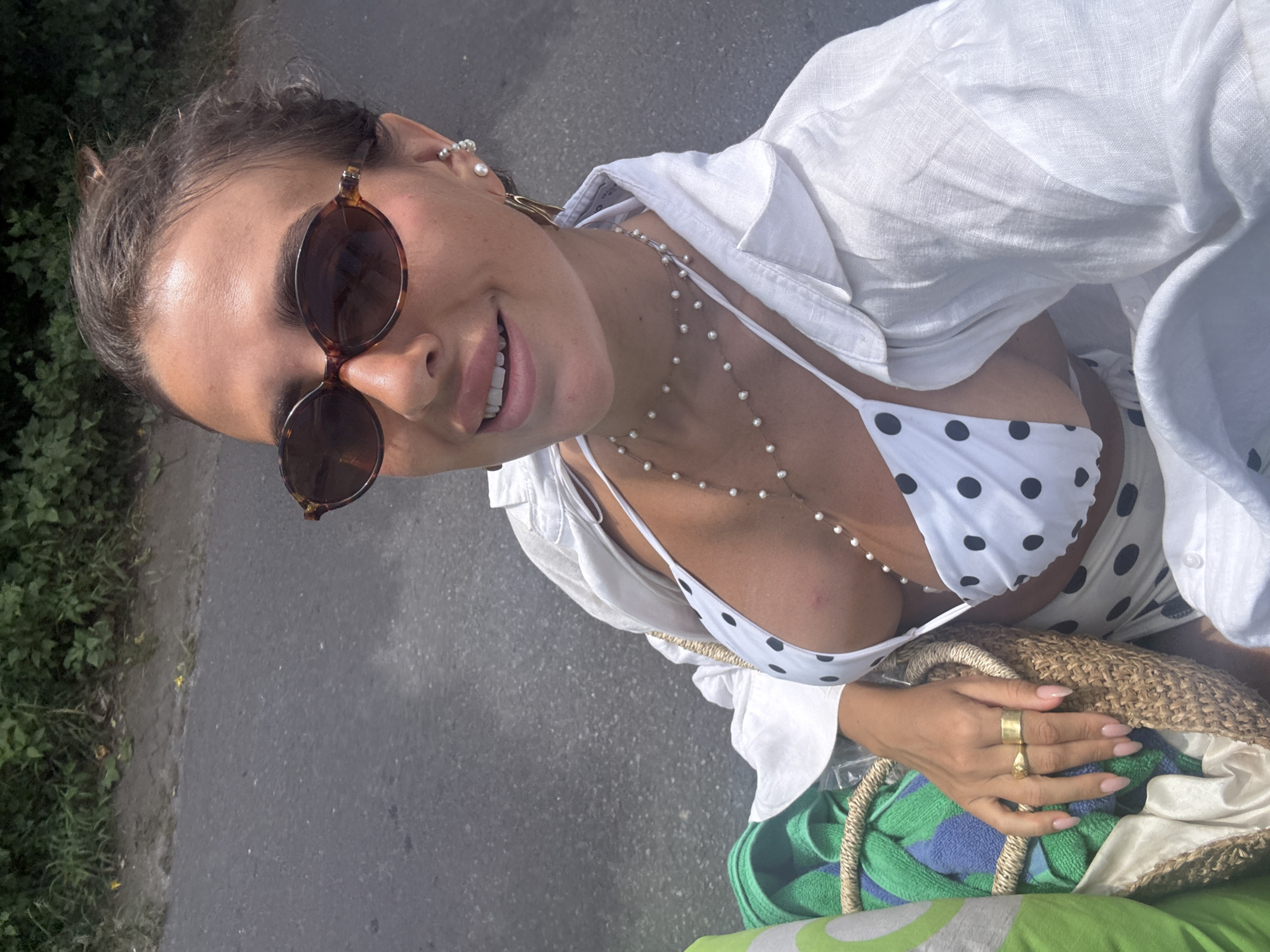 A woman is seen from a high angle, smiling and looking at the camera. She is wearing sunglasses, a white polka dot bikini top under a white button-down shirt, and beaded necklaces. A woven bag is held in her hand with a green and blue towel inside. The background is a street with greenery on one side. There is no information in the image to indicate the location is Kuta Selatan, Indonesia.