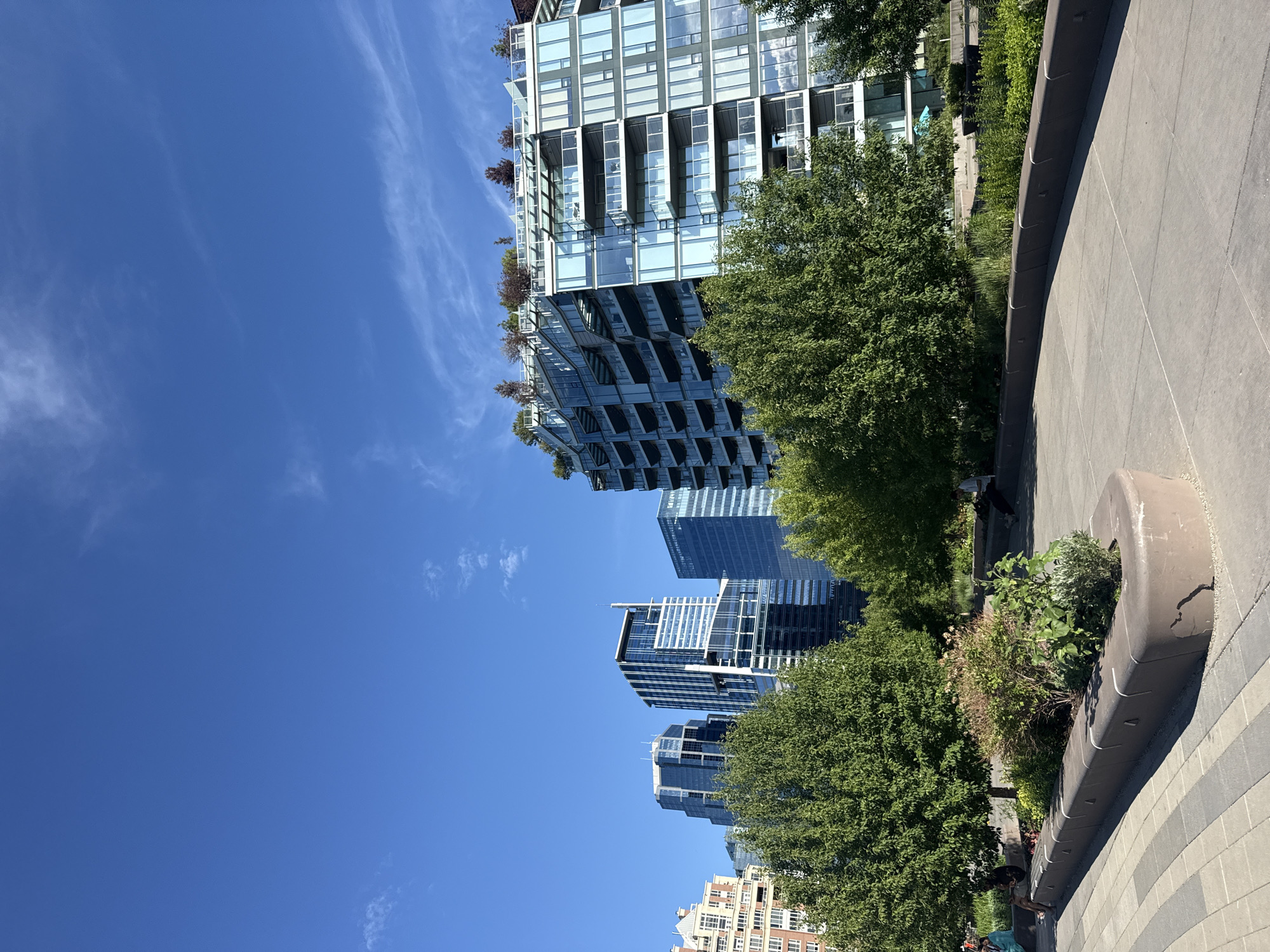 A wide-angle, eye-level, daytime shot shows a city park with a clear blue sky. In the foreground, a curved, concrete planter box with small plantings of green shrubs and flowering plants is set into a paved walkway. Beyond the planter, a paved path curves to the right. Behind the path, mature green trees with full canopies line the edge of the park. In the mid-ground and background, a cluster of modern, multi-story buildings with glass facades and balconies rise against the sky. The building on the right, a residential tower, features a stepped design with greenery on its upper levels and balconies. Other taller office buildings with reflective glass exteriors are visible to the left. A few individuals are seated on benches or walking in the park, appearing relaxed. The overall impression is of an urban green space designed for leisure and enjoyment. This scene is located in Calgary, Canada.