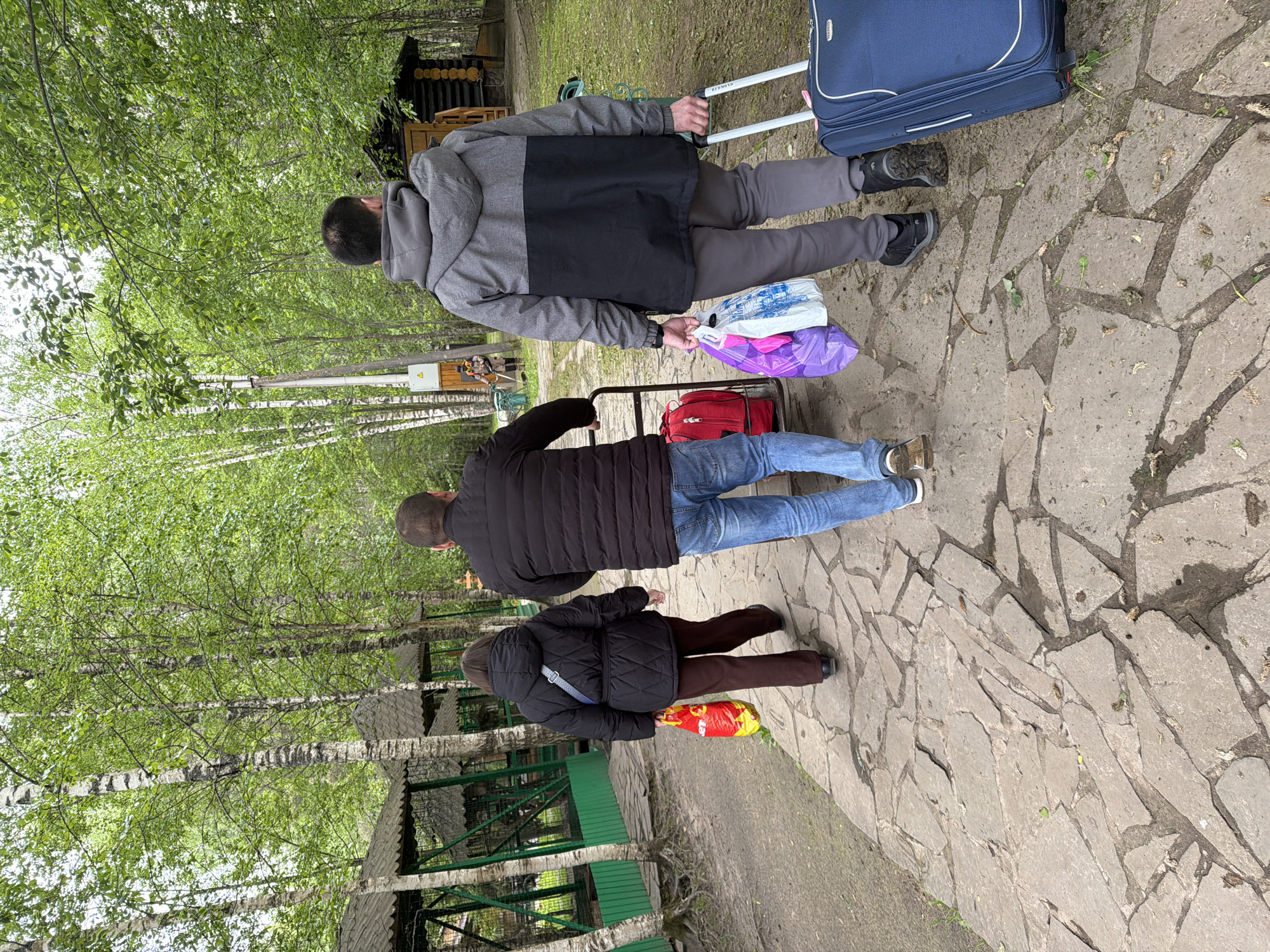 The image shows three people walking on a stone path in a park-like environment. The location is identified as Zaplav'e, Russia, based on geospatial metadata.
At the top of the image, a person is pulling a wheeled suitcase and carrying shopping bags. They are wearing a gray and black hooded jacket. Below them, a second person is pulling a wheeled cart and is wearing a black jacket and blue jeans. The third person, at the bottom of the image, is wearing a black jacket and brown pants, and they are carrying a red package.
The background consists of trees with green foliage and a green metal structure, possibly a gazebo or covered walkway. The stone path is composed of irregularly shaped stones.