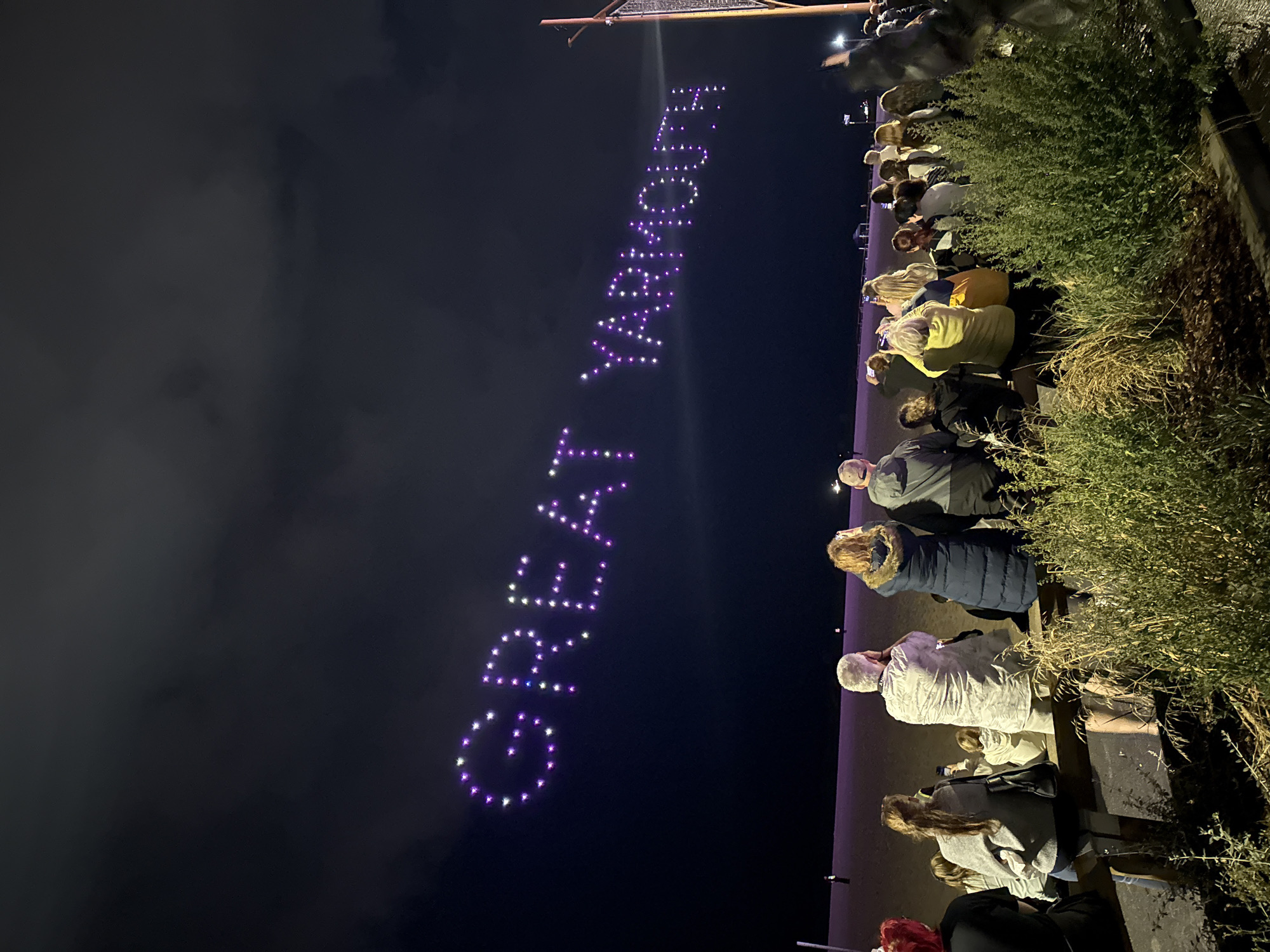 An outdoor nighttime scene depicts a crowd of people of various ages observing a drone light show in the sky above the beach in Great Yarmouth, United Kingdom. The drones, forming the words "GREAT YARMOUTH" in large, luminous purple letters, are suspended against a dark, cloudy sky. The crowd is seated and standing along a wooden barrier in the foreground, facing the illuminated text. The beach is visible in the mid-ground, with a flat, sandy expanse. The overall atmosphere is one of public spectacle and enjoyment.