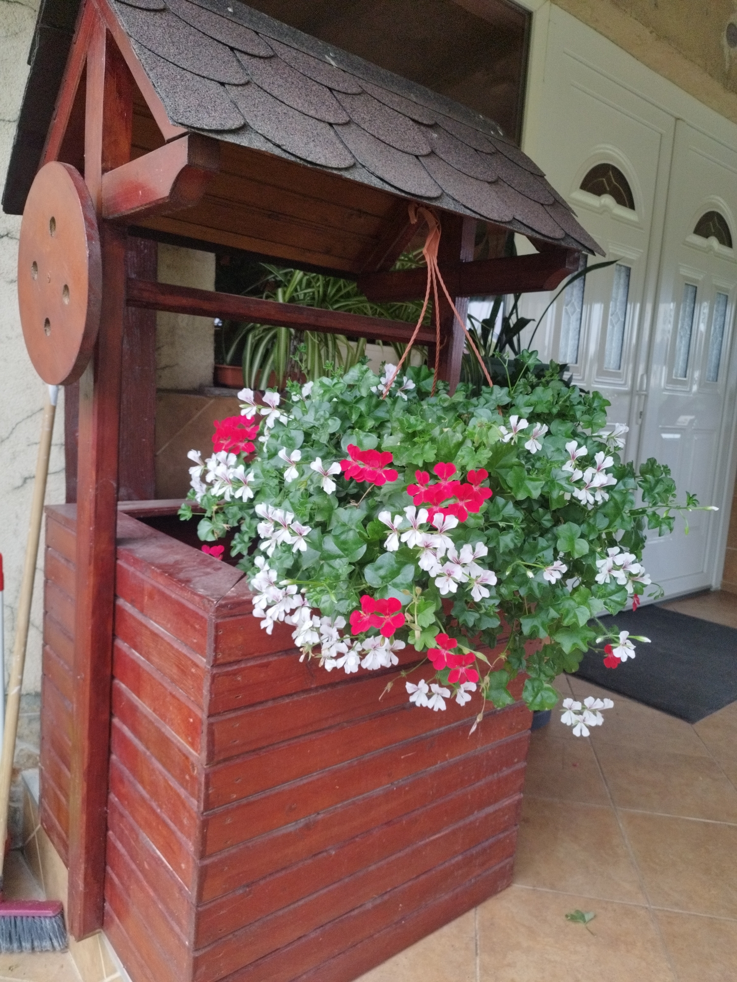 A wooden wishing well planter with a shingled roof stands on a tiled patio. The planter contains a lush arrangement of red and white geraniums. A broom leans against the side of the wishing well. In the background, a white double door with arched glass panels is visible. The scene appears to be located on the exterior of a residential building.