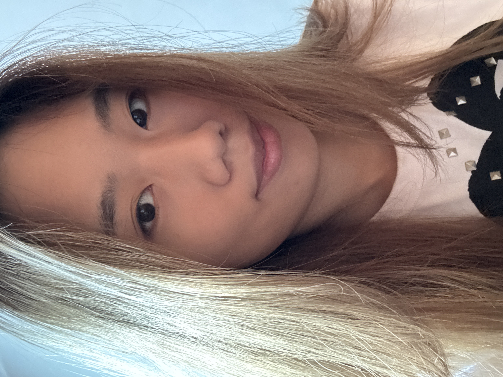 This is a close-up, selfie-style portrait of a young East Asian woman with long, straight, honey-blonde hair that falls across her face. She has dark eyes, thin eyebrows, and a neutral expression with a slight smile. She is wearing a white t-shirt with a black graphic design embellished with small silver studs. The background is a soft, light blue. There is no indication of the location being Guandu, China.