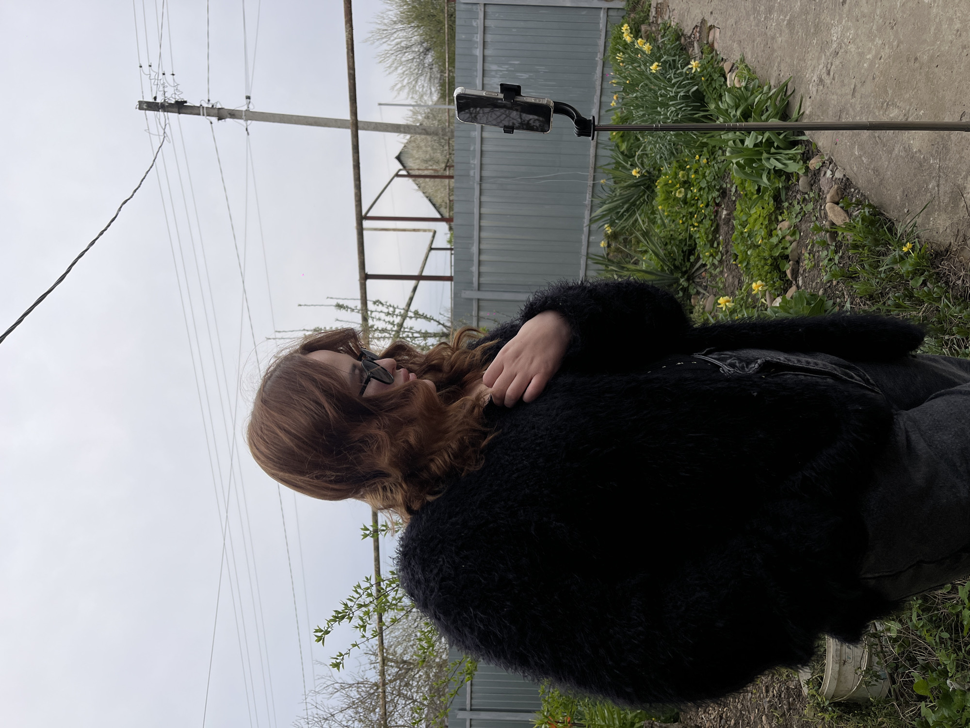 A woman with long red hair and dark sunglasses is standing in a yard, wearing a fuzzy black jacket and dark pants. She has one hand near her neck. Next to her is a black smartphone attached to a stand. Behind her is a fence, a small building with a triangular roof, a utility pole with wires, and flowers growing in a garden bed. The location is Novopokrovskaya, Russia. The sky is overcast.