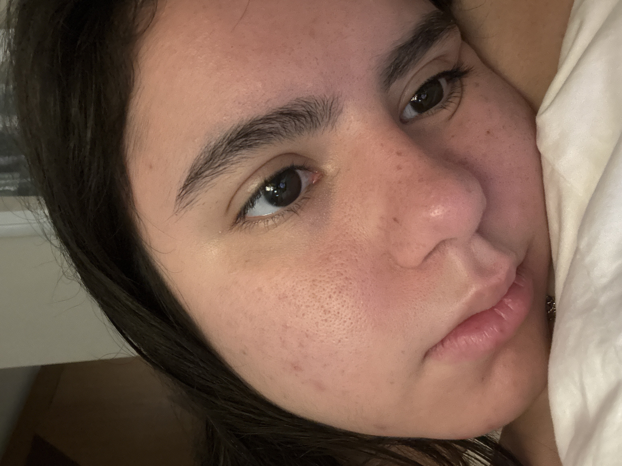 A close-up, high-angle view depicts a person's face resting against a white pillow. The individual has dark hair, prominent eyebrows, brown eyes, and a fair complexion with visible pores and some minor skin imperfections. The face fills most of the frame, with a portion of the white pillow visible on the right side. The background appears to be a dimly lit interior space. The person's expression is neutral. The location cannot be determined from this image alone.