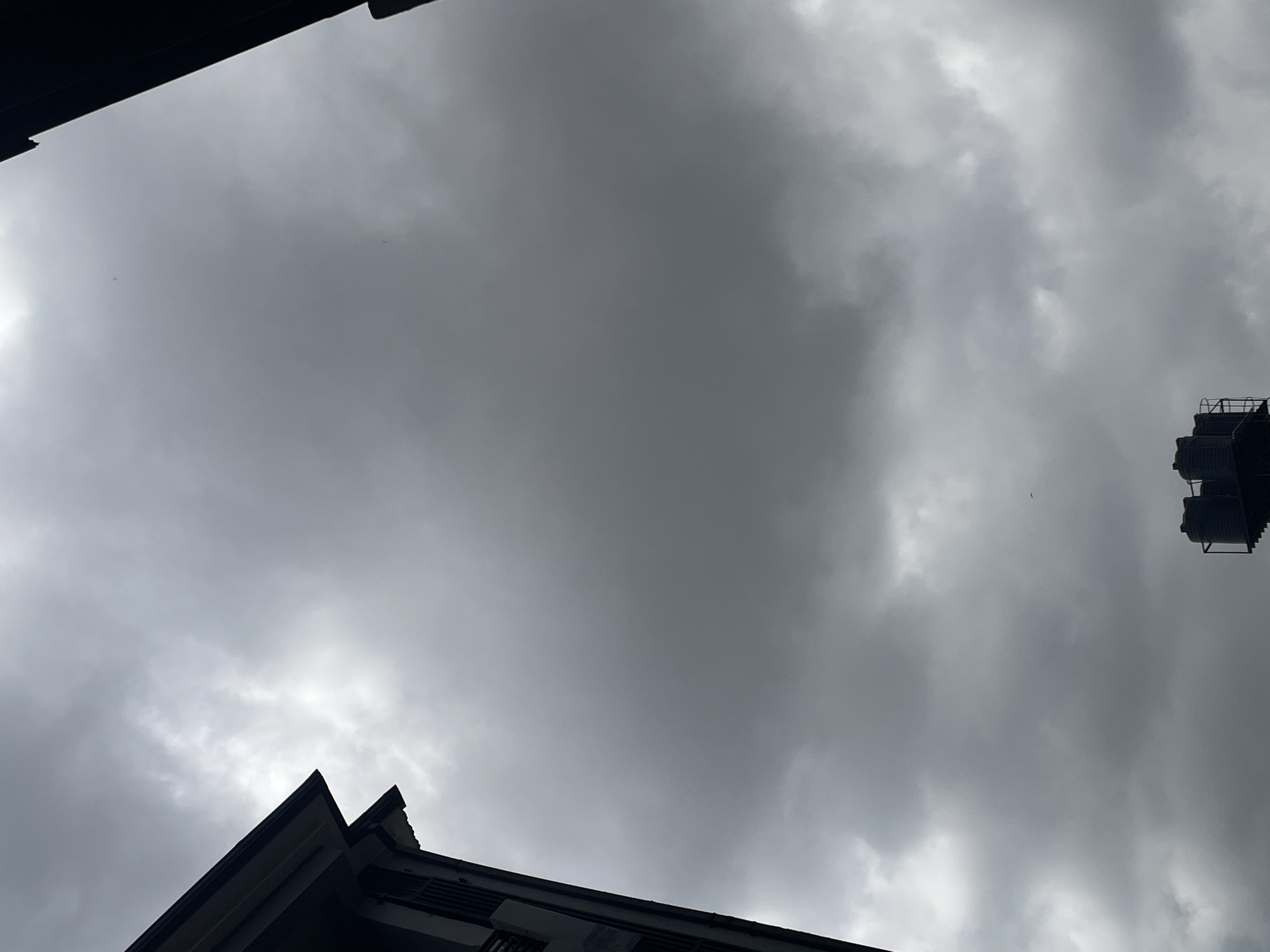 This media file captures a view of a cloudy sky from a low angle, suggesting it was taken from the ground looking upwards. The predominant feature is the overcast sky, filled with dense, dark gray clouds, indicating a possibly stormy or overcast day. The diffused light suggests it is likely daytime, but the cloud cover obscures any direct sunlight.
In the foreground, the dark silhouettes of building rooftops are visible. One rooftop in the lower left corner has a sharp, angular design, indicative of modern architecture. Another rooftop feature, possibly a water tank or similar structure, is visible in the upper right portion of the image, also silhouetted against the sky. These structures suggest an urban or semi-urban setting, consistent with the provided location context of Abuja, Nigeria.
There are no people visible in the frame. The overall mood conveyed by the image is somber and somewhat dramatic due to the heavy cloud cover. The lack of clear sunlight and the dark tones of the buildings contribute to this atmosphere. No text is visible in the image. The scene depicts a moment of a cloudy day, with architectural elements framing the sky.