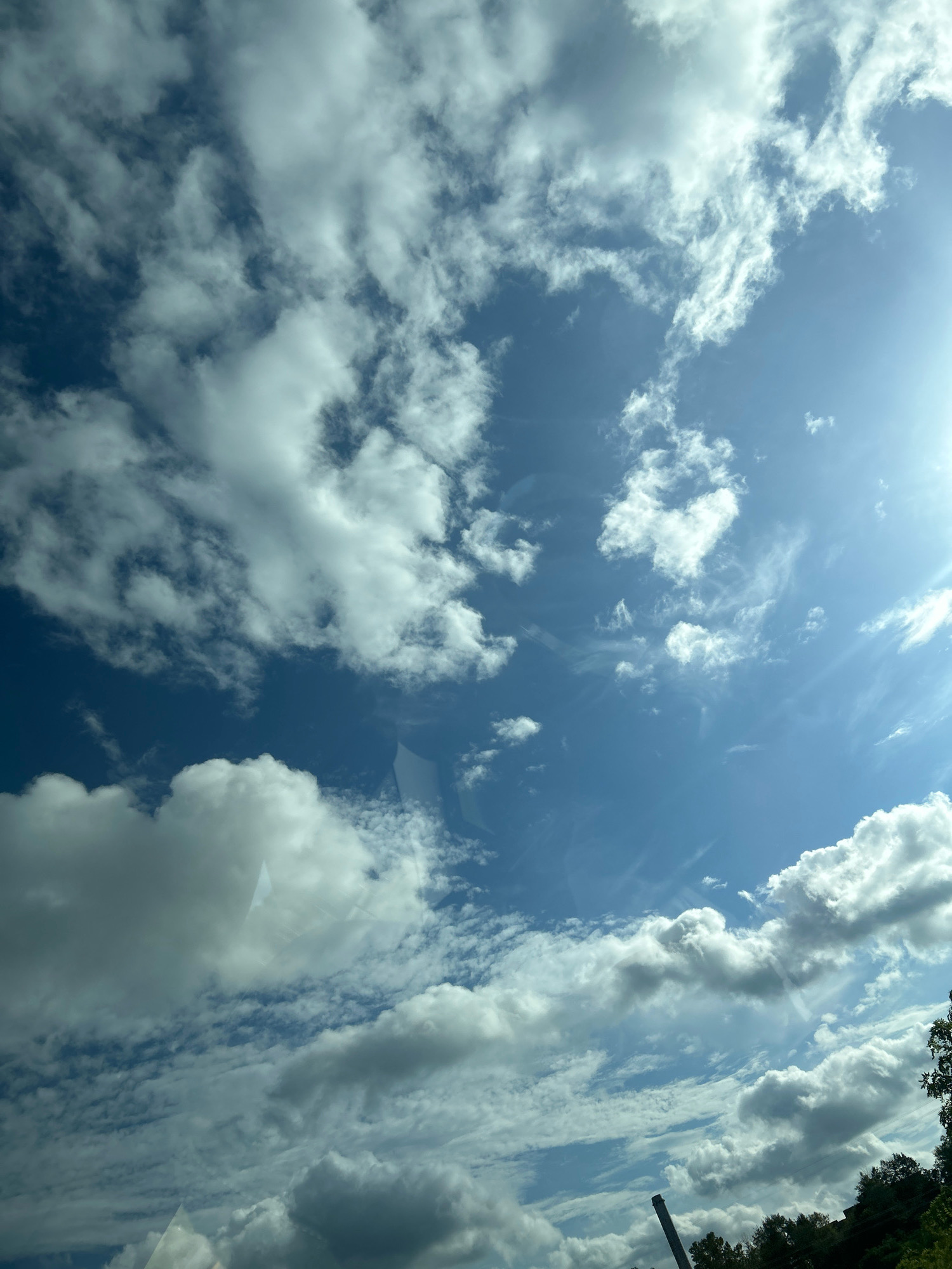 The image is a low-angle shot of a bright blue sky filled with a variety of clouds. Some clouds are large and puffy, while others are wispy and thin. The sun is visible in the upper right corner of the image, casting a bright light. The bottom of the image shows a dark, silhouetted line of trees and what appears to be a tall industrial smokestack or tower. The image is taken from inside a vehicle, as indicated by reflections on the surface of what seems to be a window.
The scene is consistent with the environment of Collier Township, United States of America, given the presence of trees and industrial structures that could be found in such a suburban or semi-industrial area. However, there are no identifiable landmarks or specific details that would pinpoint the exact location within Collier Township.
There are no people visible in the image. The subjects are the sky, clouds, sunlight, trees, and the industrial structure. The action is static, capturing a moment in time. There are no interactions depicted. The environment is outdoors, under a partly cloudy sky on a sunny day.