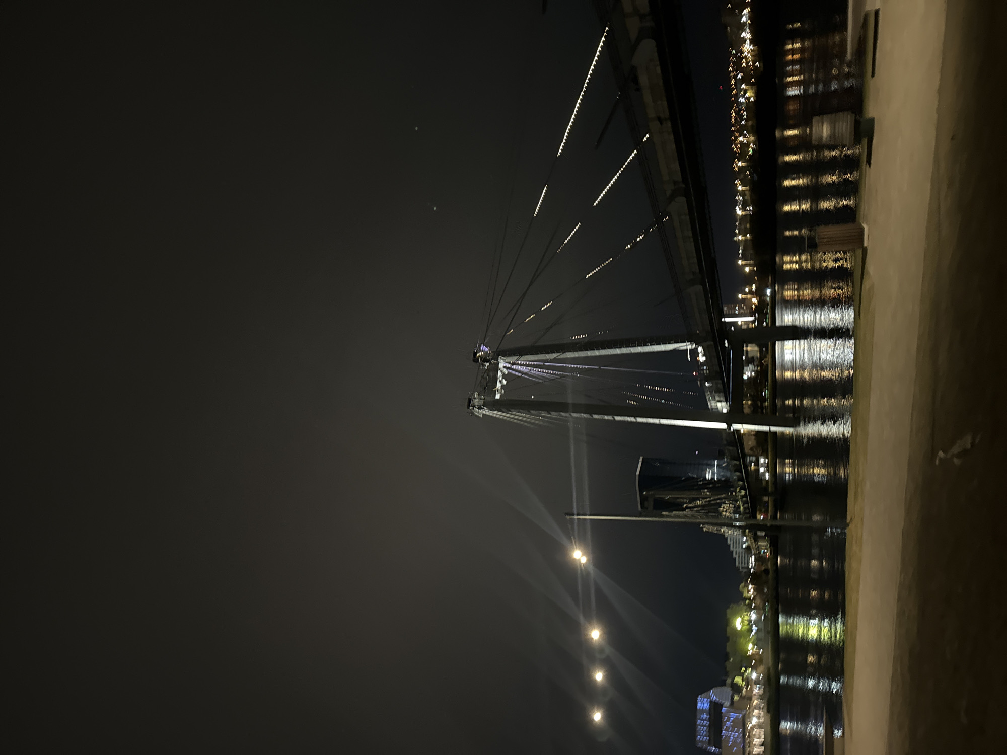 The image depicts a nighttime scene featuring a cable-stayed bridge. The bridge is illuminated with strings of lights, creating a distinct linear pattern against the dark sky. The structure's tower is visible, with cables extending from it towards the bridge deck. The river reflects the lights from the bridge and nearby buildings, creating a shimmering effect. The location is most likely the Vinogradovsky Bridge in Krasnoyarsk, Russia, judging by the bridge's structure and the presence of a river with illuminated buildings along its banks.