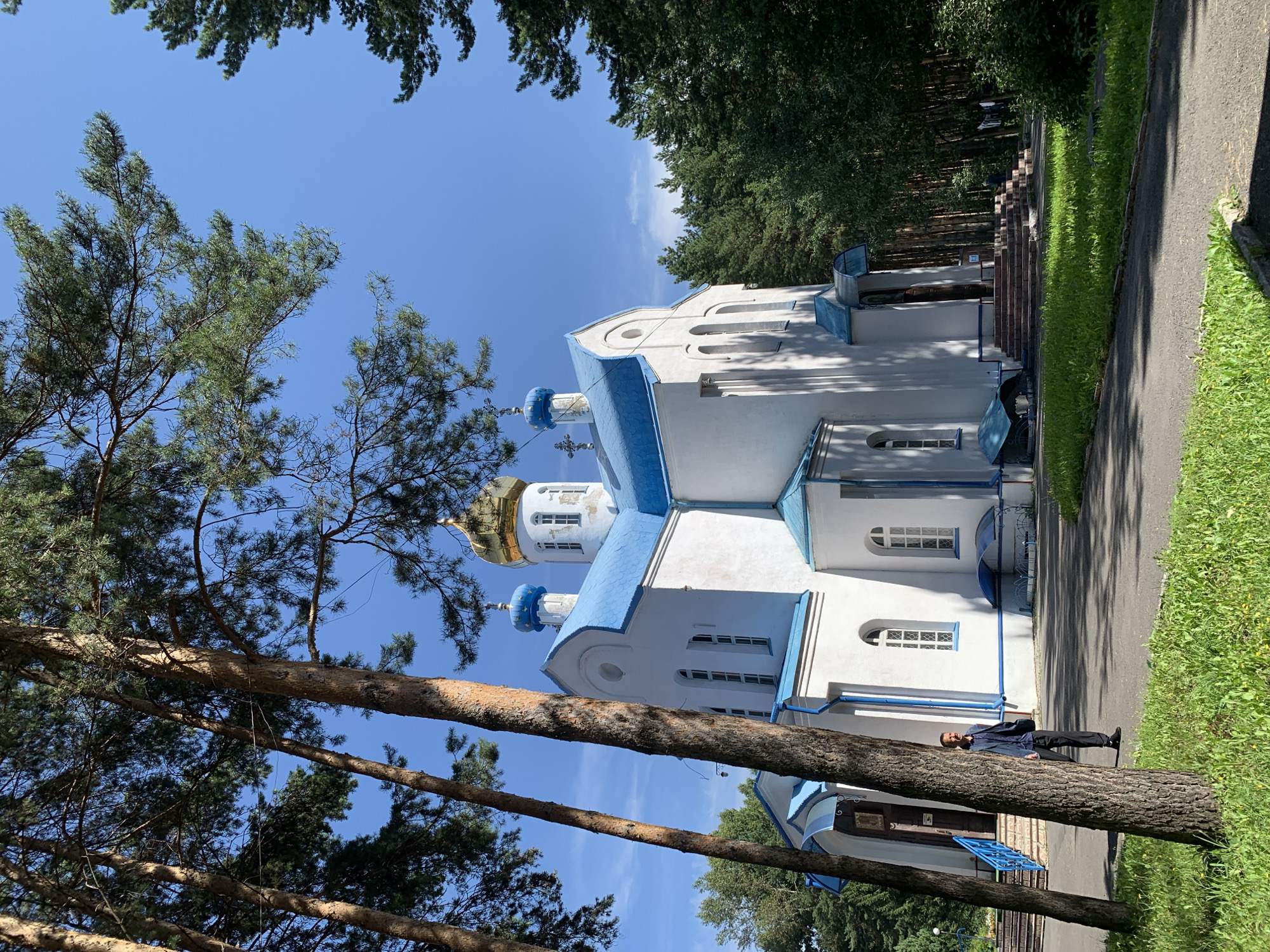 The image shows a white Orthodox church with blue roofs and a golden dome, set amidst a cluster of tall pine trees under a clear blue sky. A paved pathway leads to the church entrance, with a grassy area to the left. A lone man in a dark blue shirt and dark pants stands on the pathway, facing away from the camera and looking towards the church. He is positioned to the left of the pathway, partially obscured by a large pine tree trunk. The church architecture is visible, featuring arched windows and smaller blue domes. The overall environment suggests a serene, possibly rural or park-like setting within or near the city of Prokopyevsk, Russia.