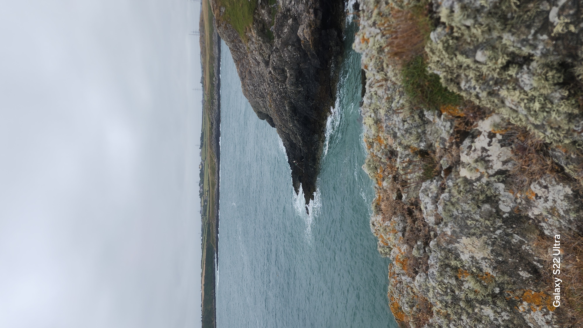 A view of the coast near Cemaes, United Kingdom. The image features a rocky shoreline with the sea to the left and a distant landmass with some buildings. The sky is overcast. In the foreground, rocks are covered with lichen. The water is choppy. There is no discernable human activity or interaction in the scene. The overall impression is a natural, coastal landscape under a cloudy sky.