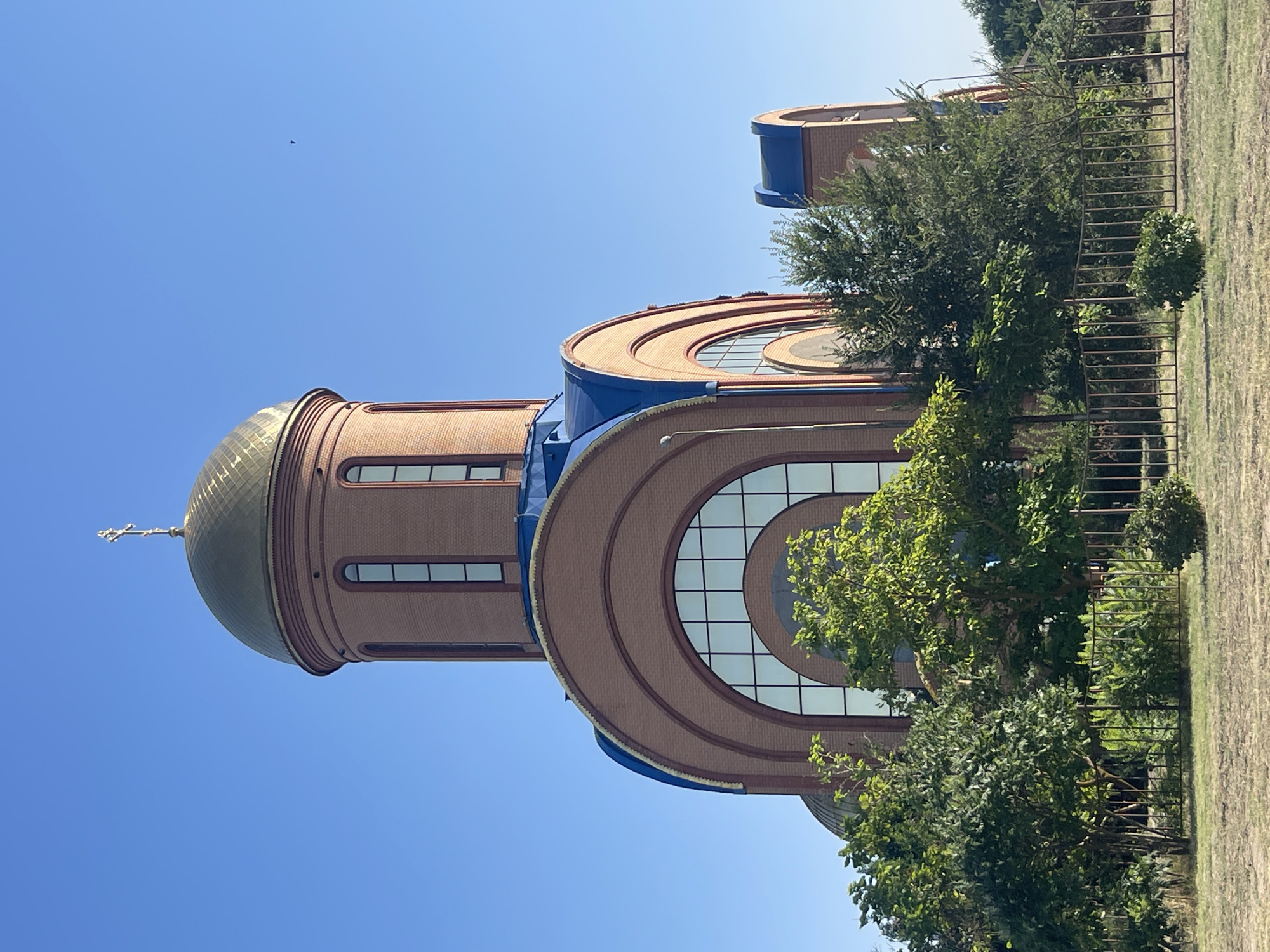 This is a photograph of the Holy Trinity Cathedral in Budyonnovsk, Russia. The cathedral is constructed of red brick with a golden dome topped with a cross. The dome is surrounded by a blue roof. Large arched windows with multiple panes are visible on the cathedral. Green trees and bushes are in the foreground, and a metal fence runs along the edge of the property. The sky is clear and blue.
