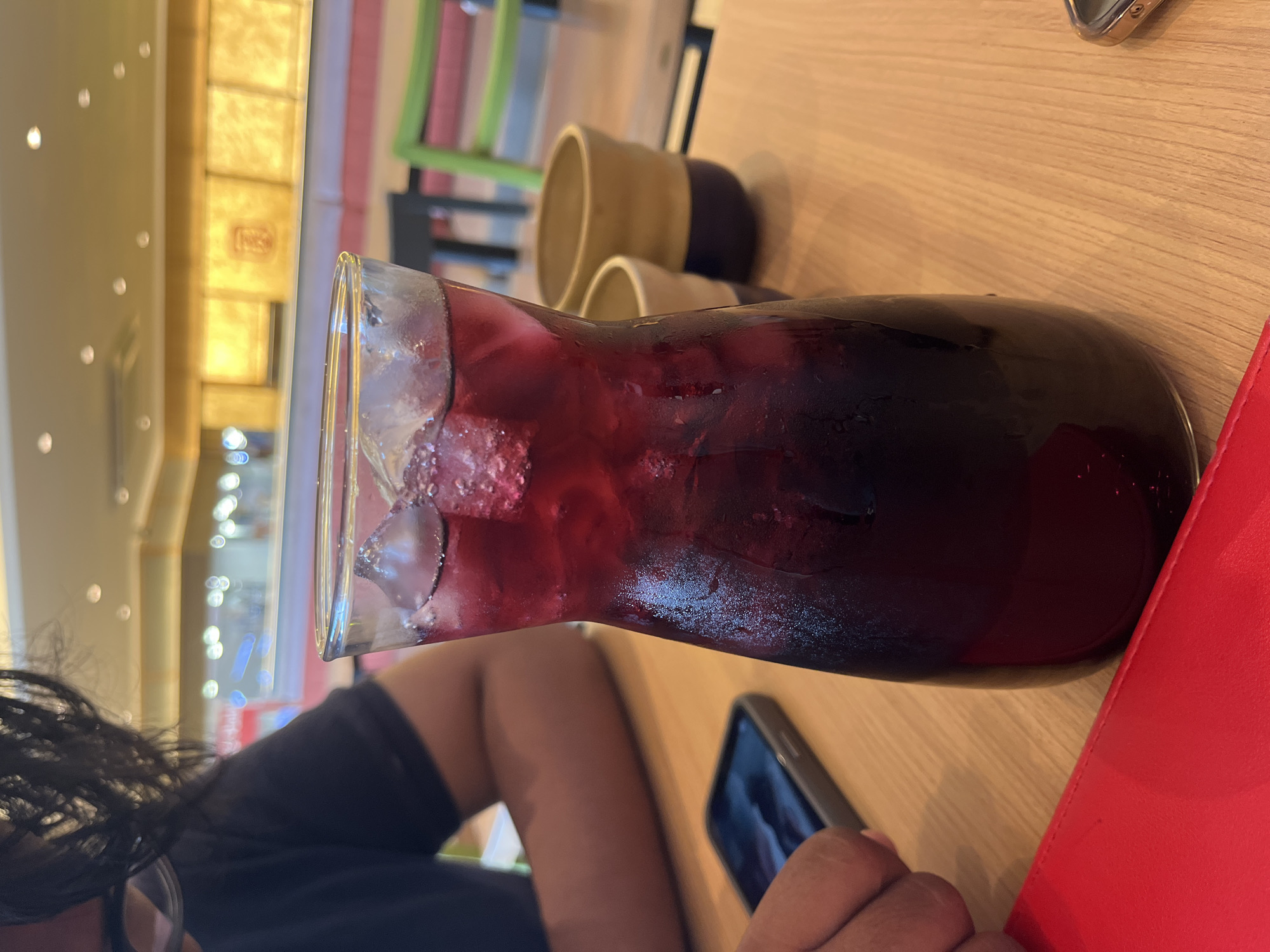 The image shows a pitcher of dark red liquid, possibly juice or sangria, filled with ice. The pitcher sits on a light-colored wooden table next to a smartphone and a red object, possibly a book or wallet. Two ceramic cups are visible behind the pitcher. A person with dark hair is partially visible in the lower left corner. The background shows a brightly lit interior space, likely a restaurant or cafe, with colorful decorations. It is not possible to verify the location is Lhokseumawe, Indonesia from the image alone.
