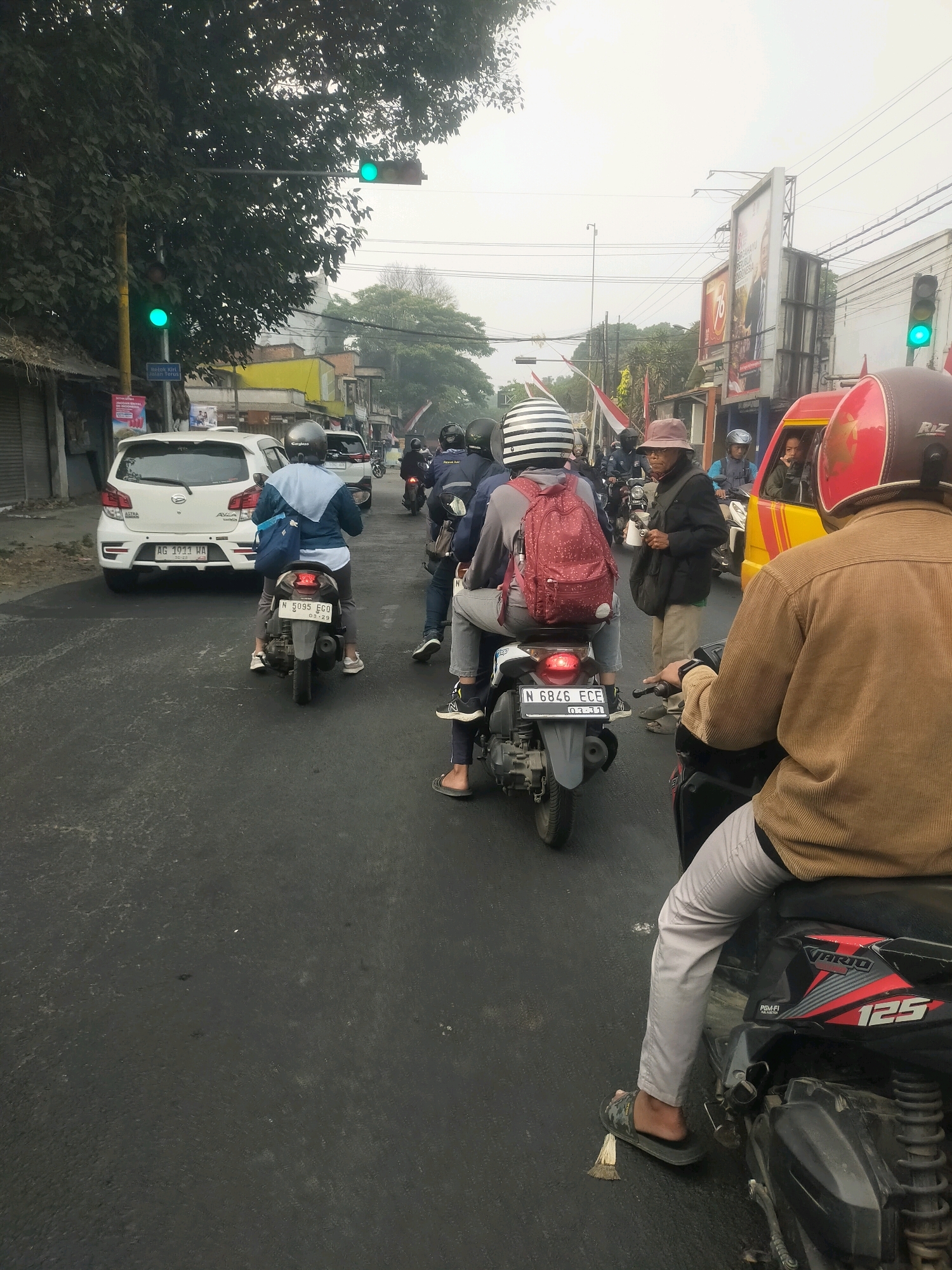 The image depicts a street scene in Gading, Indonesia, likely during daylight hours due to the visible traffic lights and general illumination. The scene is characterized by a moderate amount of traffic, primarily consisting of motorcycles and a few cars. The road surface appears to be asphalt, showing signs of wear and tear, with some scattered debris.
In the foreground, a person is riding a motorcycle, facing away from the camera. This individual is wearing a tan corduroy jacket and light-colored pants, with a red helmet. Their right foot is resting on the ground, wearing a sandal. The motorcycle is black with red and white accents and displays the number "125".
Behind this motorcycle, other vehicles are visible. One motorcycle carries two people, the rider in front wearing a striped helmet and a grey backpack, and a passenger behind. Another motorcycle is ridden by a person wearing a blue jacket and a dark helmet. Further down the road, a white car is prominent, with a license plate that reads "AG 1911 WA". To the right of this car, another white car is visible.
The background shows buildings lining the street, with various signage. A large billboard is visible in the distance, featuring Indonesian text and images. Trees with dense foliage are present on the left side of the road, casting shadows. Traffic lights are visible at an intersection, with at least one green light illuminated. The sky is overcast, contributing to a diffused light condition. The overall impression is one of a busy urban street environment.