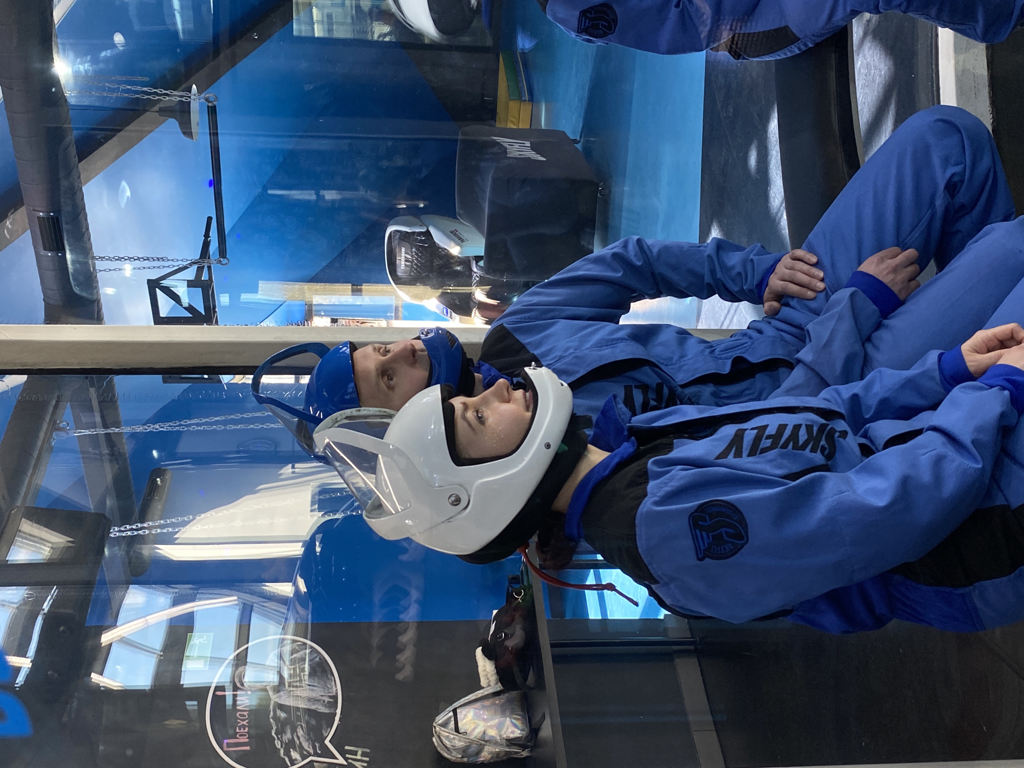 Two people in blue skydiving suits and helmets are preparing for an indoor skydiving experience at the Aerotruba FlyStation in Izhevsk, Russia. The individuals are wearing full-face helmets, and one is wearing a white helmet. The backdrop reveals the structure of the indoor skydiving facility, with glass walls and support beams.