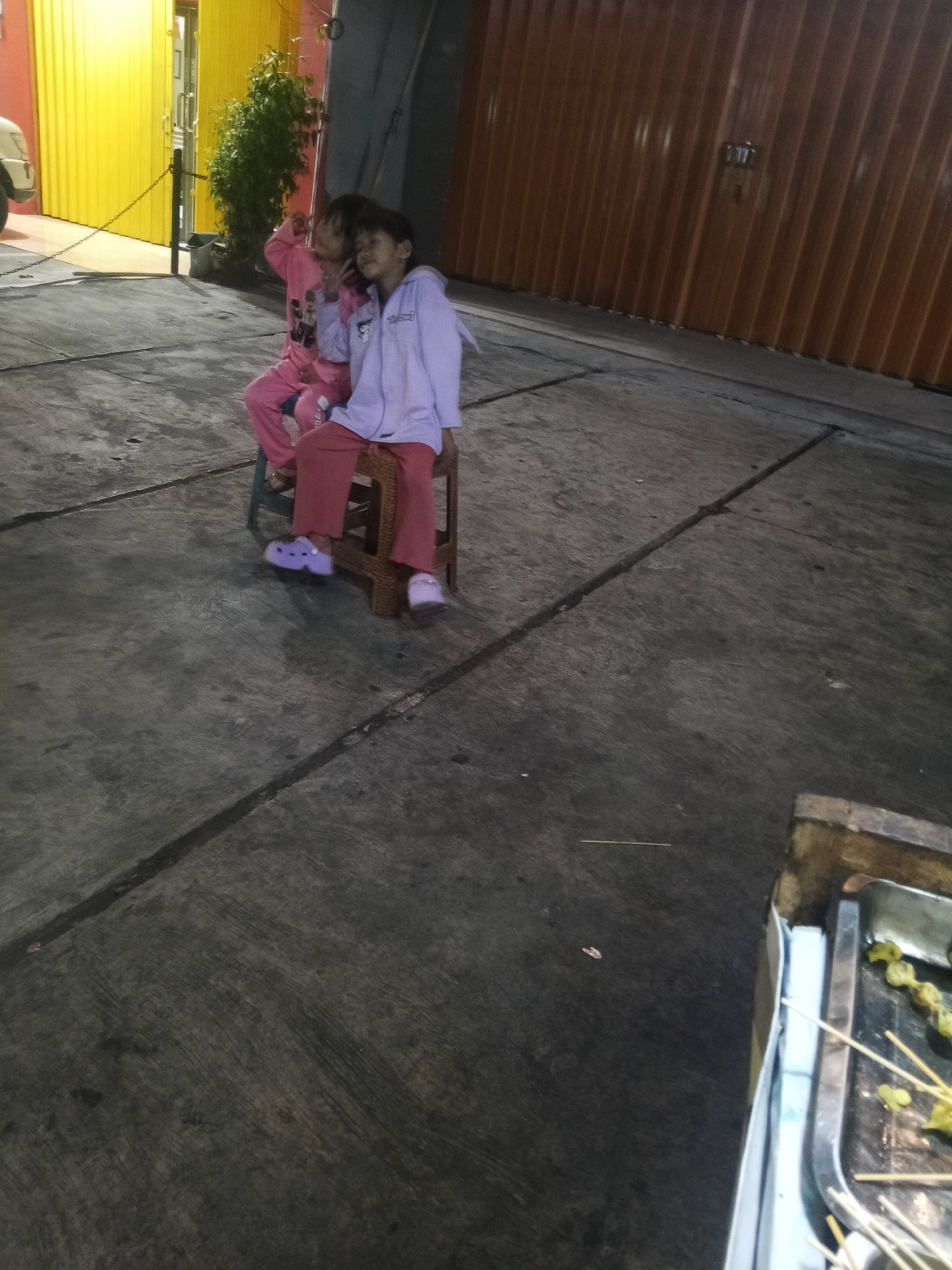 Two young girls, one in a pink outfit and the other in a purple jacket and pink pants, sit on chairs outdoors. The girl in pink is slightly behind and to the left of the girl in purple, who has her head tilted down. They are positioned on a concrete surface, with a yellow wall and a closed orange corrugated metal door in the background. To the lower right of the image, a metal tray with skewers of food is visible, suggesting a food stall or preparation area. The lighting indicates it is nighttime. This scene is located in the city of Lubuklinggau, Indonesia, likely in a public or semi-public outdoor space such as a street or alleyway adjacent to commercial buildings.