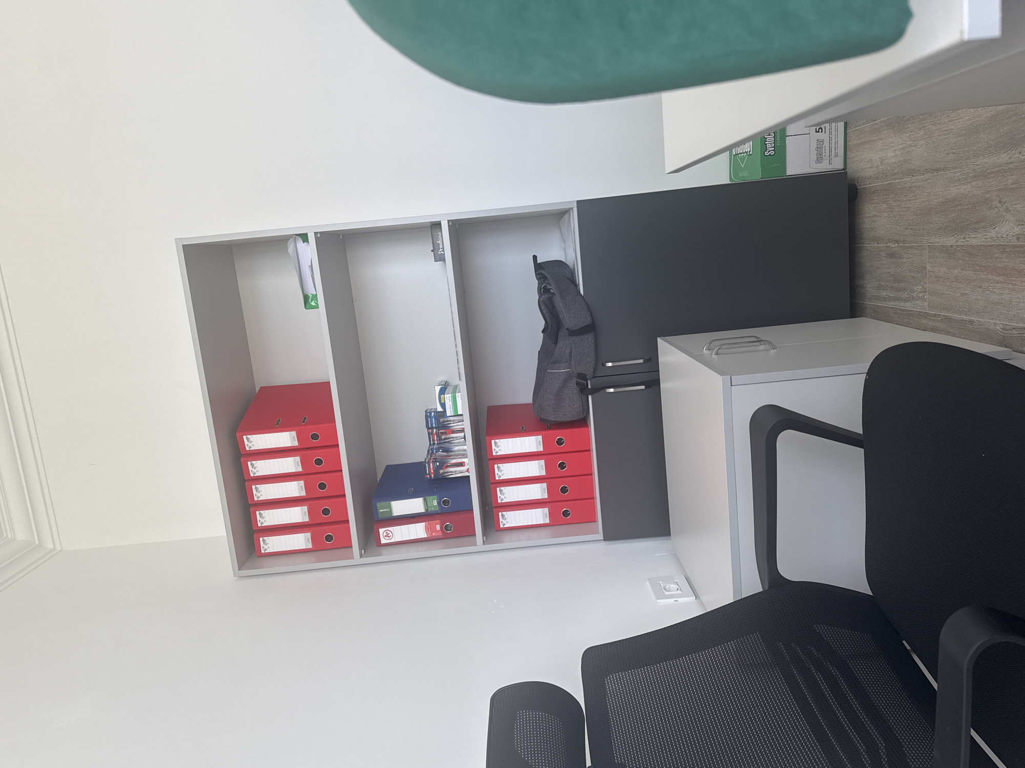 In an interior scene, likely an office, located in Sabunchu Raion, Azerbaijan, a black mesh office chair with black armrests is positioned to the left of a white desk with a grey drawer unit. The desk unit has visible handles and is placed adjacent to a tall, grey shelving unit. The shelving unit contains several red binders on the top two shelves, a blue and red binder on the second shelf, and a grey duffel bag on the bottom shelf. There are also various office supplies and papers visible on the shelves. To the right of the shelving unit, a portion of a white desk with a green curved object on it is visible. The flooring consists of light-colored wooden planks. The walls are painted white, and there is a white crown molding at the top of the wall. A white electrical outlet is mounted on the wall behind the chair. A green box labeled "SvetoCopy" is partially visible on the floor to the right of the grey drawer unit.