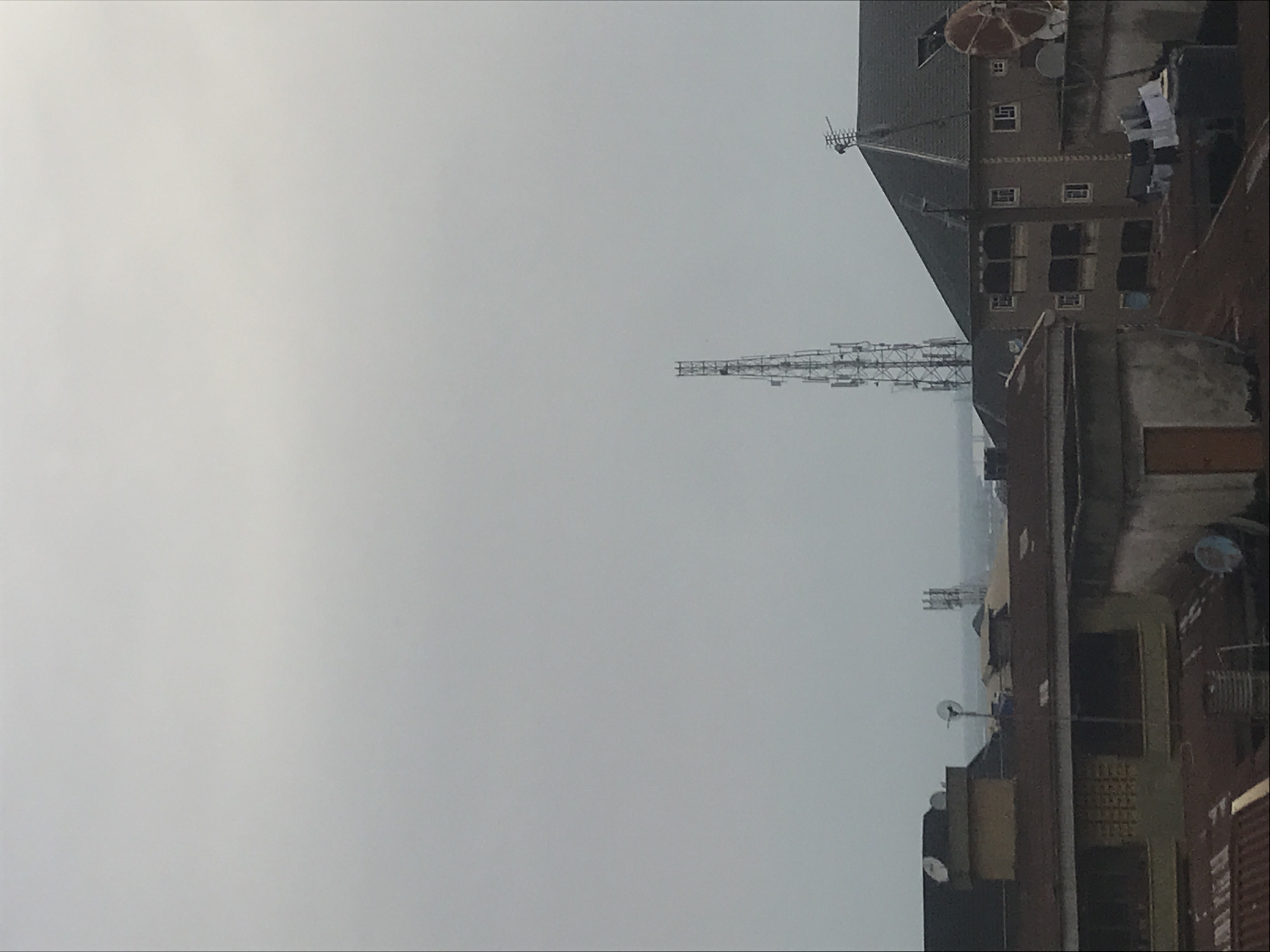 The image displays an urban or semi-urban landscape, likely in Onitsha, Nigeria, viewed from an elevated perspective. The scene is dominated by a vast, light grey, hazy sky that covers the left half of the frame and extends into the distance, suggesting an overcast day or atmospheric haze.
On the right side of the image, a cluster of buildings is visible. A prominent, multi-story building features a dark, sloped roof and multiple rectangular windows. On its rooftop, a small, patterned shade structure or vent is visible alongside a white satellite dish, and laundry is hung out to dry. Closer to the foreground, another concrete building with a flat roof is partially visible, also featuring a satellite dish. Several other structures and rooftops recede into the hazy background.
In the middle distance, standing tall against the light sky, is a slender, grey metallic lattice tower, likely a telecommunications mast, equipped with several platforms and antenna arrays. The overall impression is that of a bustling city, with typical urban infrastructure like communication towers and satellite dishes integrated into the residential and commercial areas. No people are visible, and no specific activities are taking place. There is no discernible text in the image. The time of day appears to be daytime, though the sun is obscured by the overcast conditions.