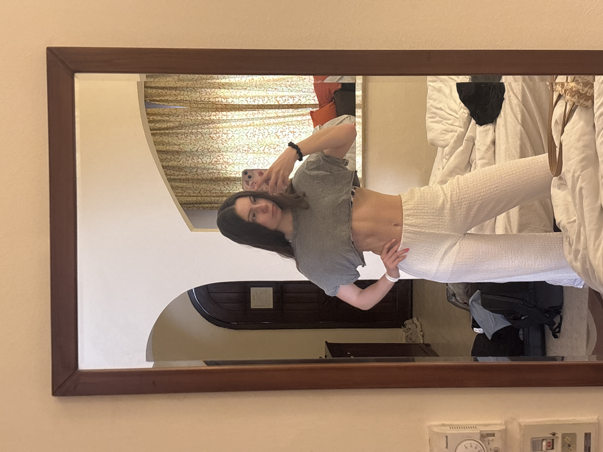 A woman is taking a mirror selfie in a hotel room in Sharm El Sheikh, Egypt. She is wearing a grey crop top and white pants. Behind her, there's a bed with white linens, patterned curtains, and a dark wooden door. A thermostat is visible on the wall below the mirror.