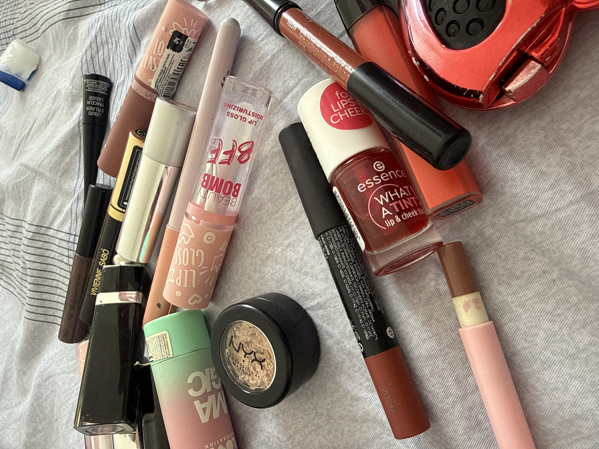 The image shows a collection of various makeup products scattered on a light gray striped surface, likely a bedsheet. The products include lipsticks, lip glosses, eyeliners, eyeshadow, and a lip and cheek tint. Brands visible are Essence, NYX Professional Makeup, Vivienne Sabó, Love Generation. Details on some products include "Lip Gloss Moisturizing," "Lasting Matte Liquid," "Liquid Eyeliner," and "What a Tint lip & cheek."
The products are not arranged in any particular order, suggesting a casual or disorganized setting. There is nothing in the image to indicate a location in Moscow, Russia.