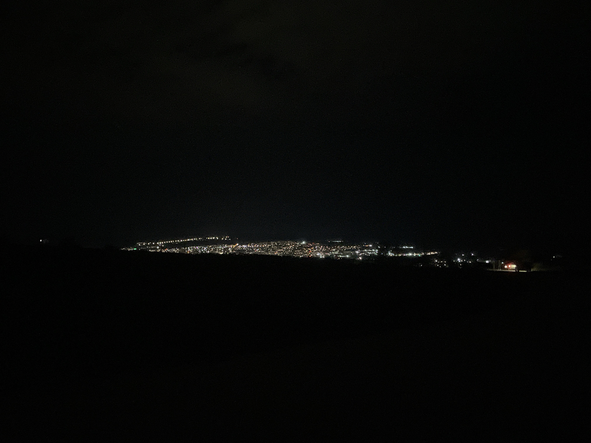 The image shows a nighttime view of the city of Razgrad, Bulgaria, from an elevated perspective. The city lights are visible in the distance, and the foreground is dark, likely representing the land or terrain from where the photo was taken. The sky is dark and mostly featureless. In the bottom right-hand corner, the lights of a vehicle, possibly a car, are visible. No people are discernible. The environment is dark and mostly unidentifiable beyond the city lights in the distance.