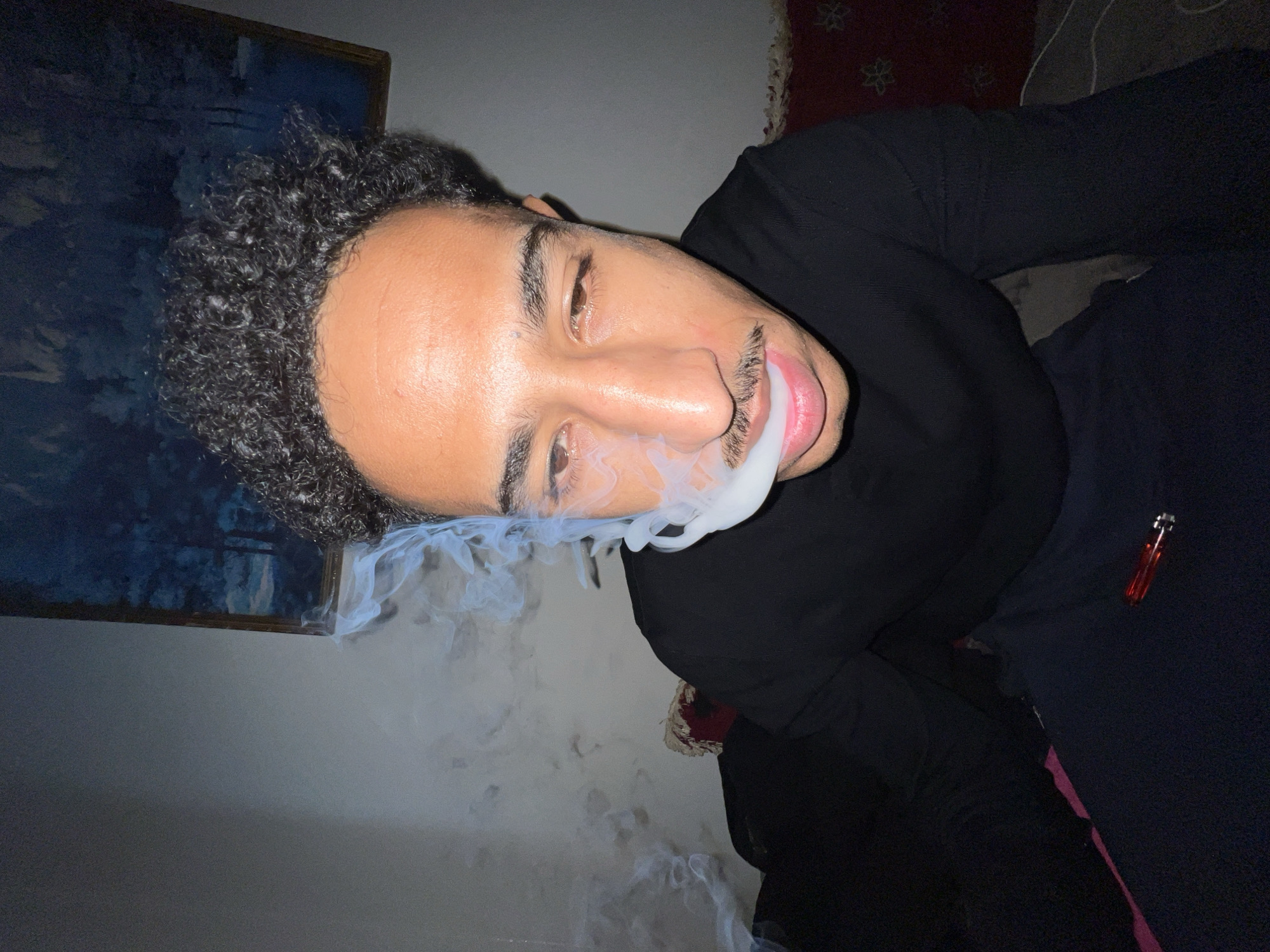 The image depicts an indoor scene in Tanger, Morocco, featuring a male subject. The subject, a young adult male with dark, curly hair and a mustache, is positioned in the foreground, facing the camera. He is wearing a black long-sleeved shirt and dark trousers. White smoke emanates from his mouth, forming a distinct ring shape before dissipating to his left. His eyes are partially closed. To the lower right of the frame, near the subject's leg, a translucent red lighter is visible on a dark fabric surface. The background consists of a light gray wall. A framed artwork, depicting a dark, abstract landscape with tree-like forms in shades of blue and black, hangs on the wall in the upper right quadrant. A dark red cushion with white embroidered patterns is partially visible behind and to the right of the subject, suggesting a seating area. The overall lighting is dim.